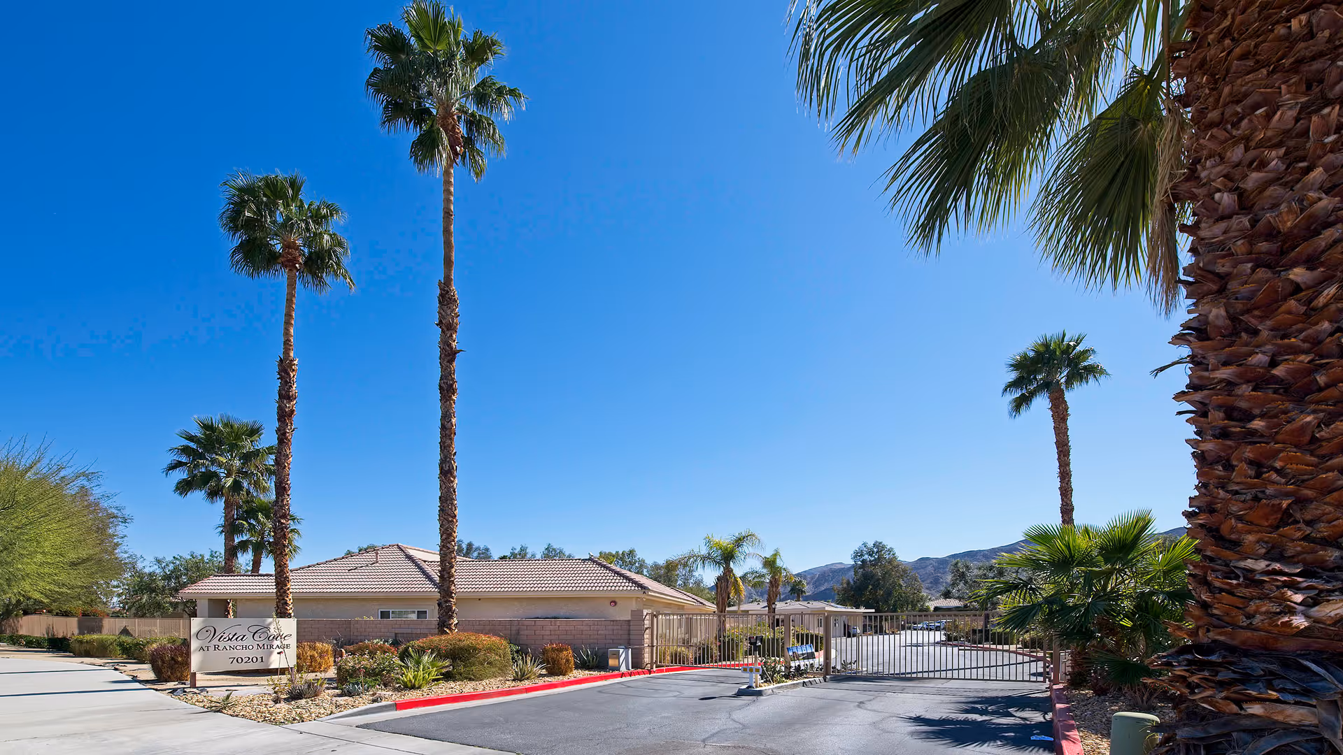 Gated entrance and driveway of Vista Cove at Rancho Mirage with palm trees, a low building, and clear blue sky.