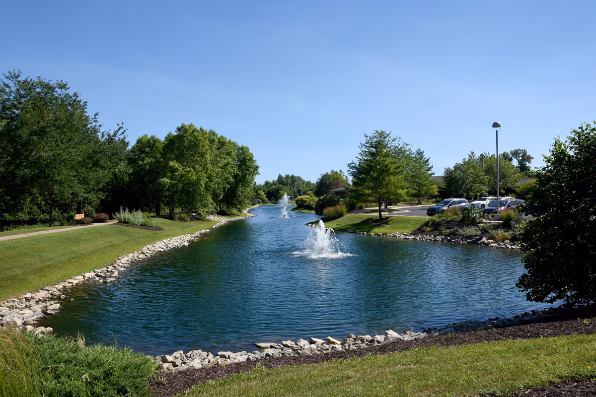 Serene pond with fountains, flanked by trees and a nearby parking lot.