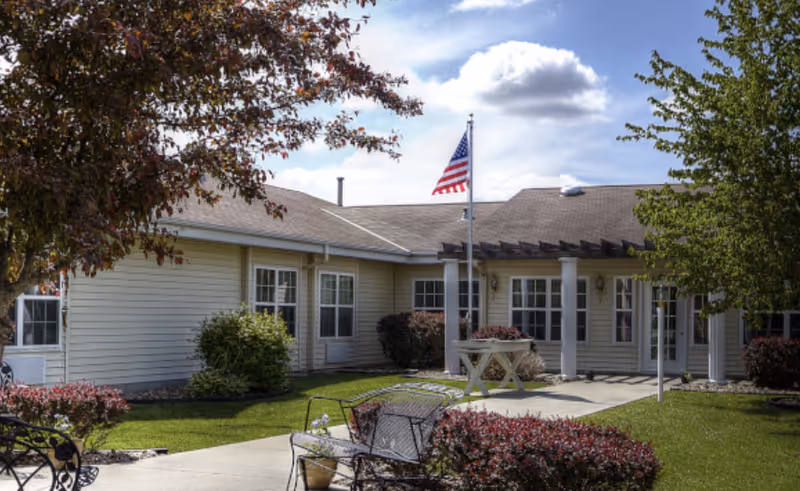 Front exterior of a single-story senior living facility with a flagpole, patio seating, walkway, and landscaped lawn.