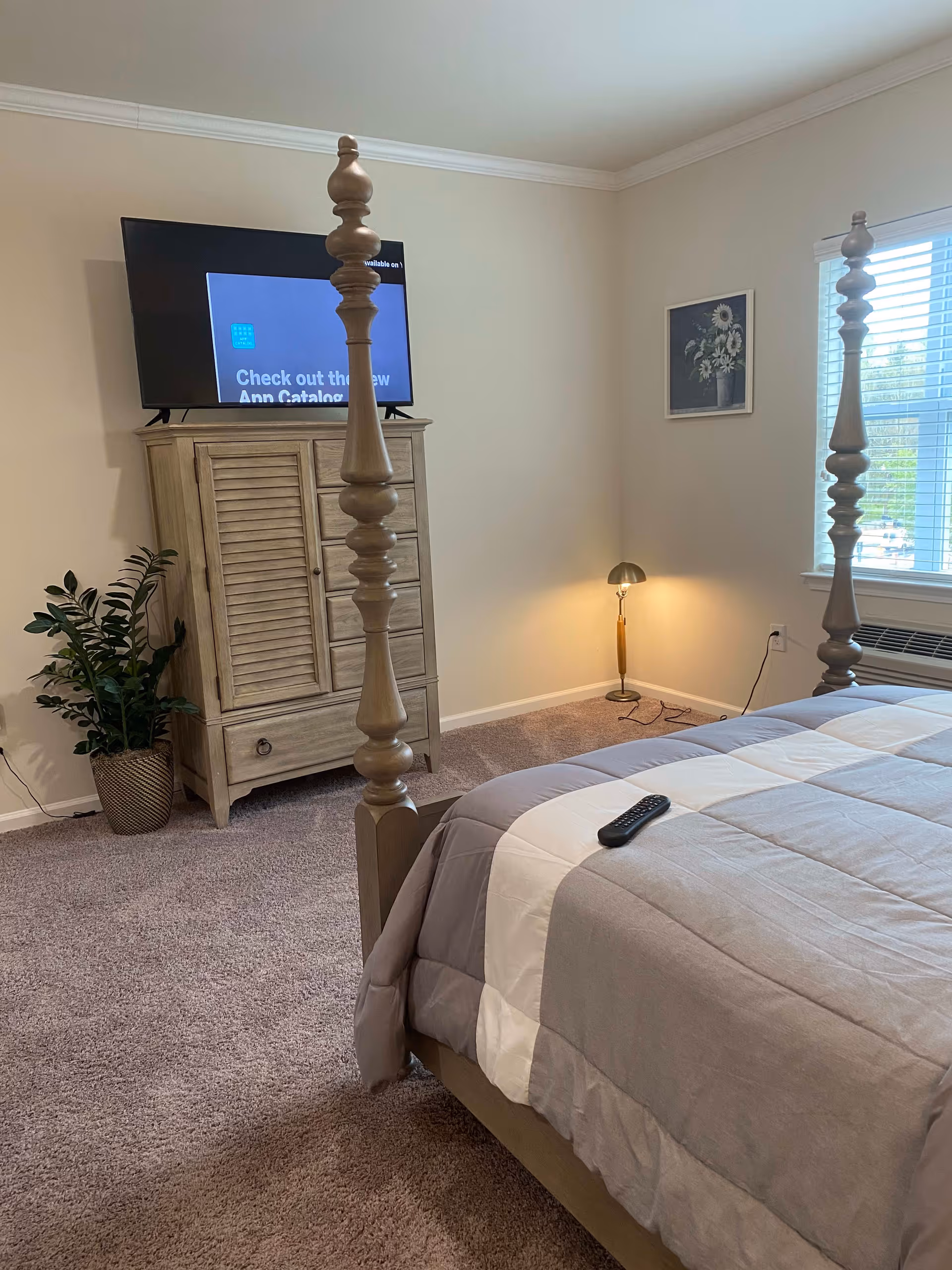 A bedroom with a wooden four-poster bed covered with a gray and white comforter. A TV is mounted on a wooden cabinet against the wall, next to a potted plant. A small lamp is on the floor near a window with blinds, and a framed floral picture hangs on the wall.