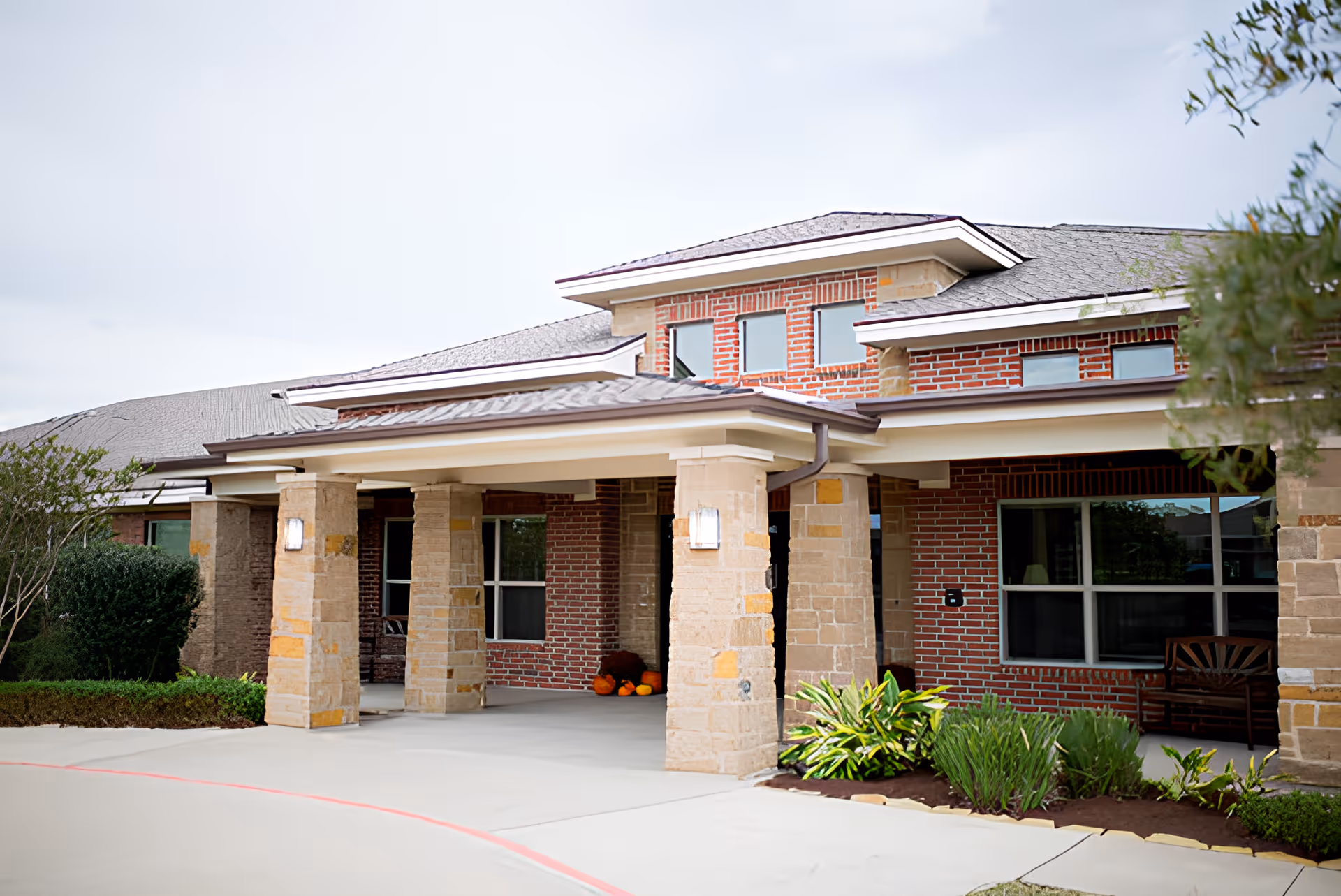 Front entrance of a brick memory care building with a covered porte-cochère supported by stone columns, windows, and landscaping.