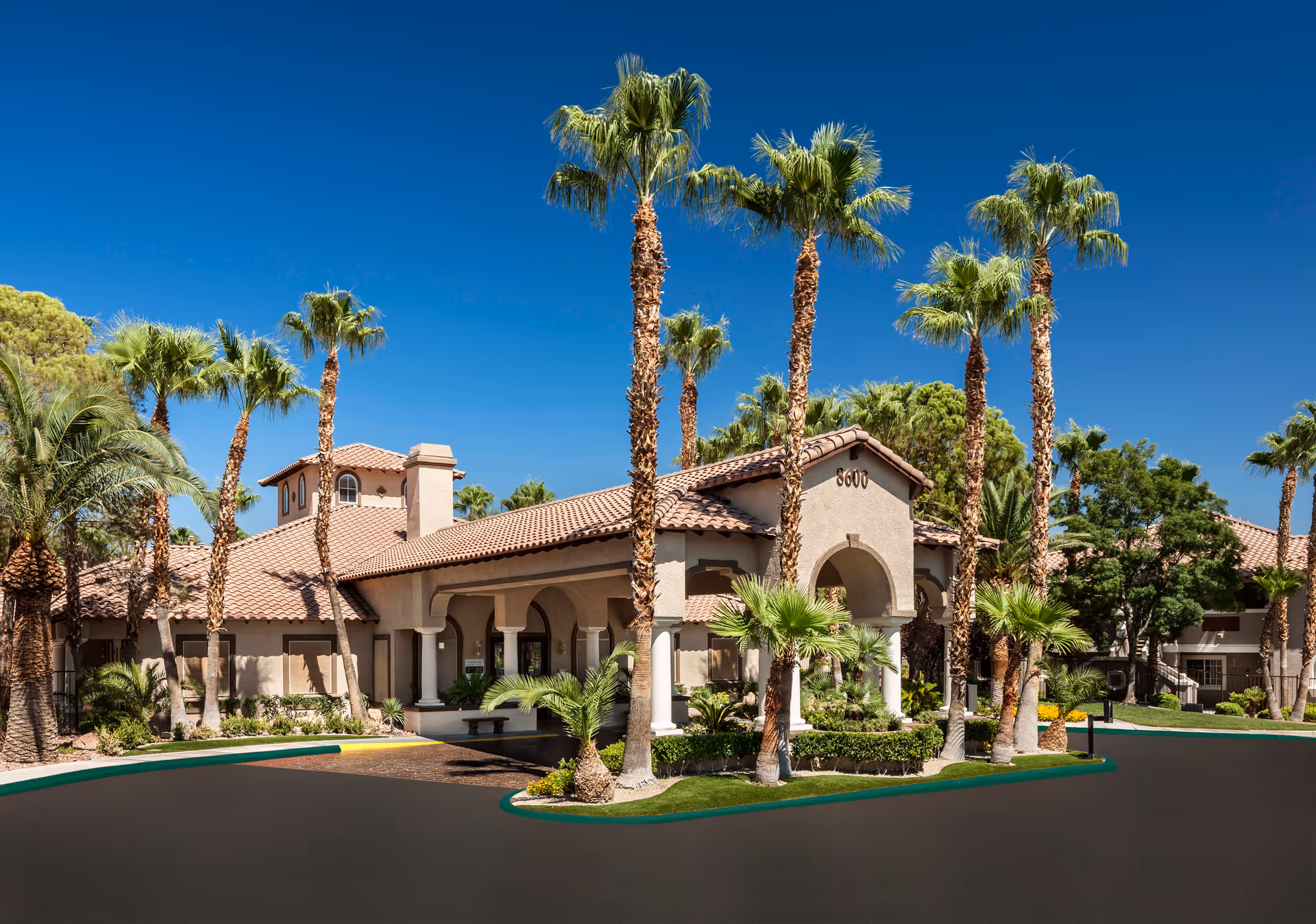 Exterior view of Destinations Pueblo facility featuring a Mediterranean-style building with a tiled roof, arched entrance, and surrounded by tall palm trees and landscaped greenery under a clear blue sky.