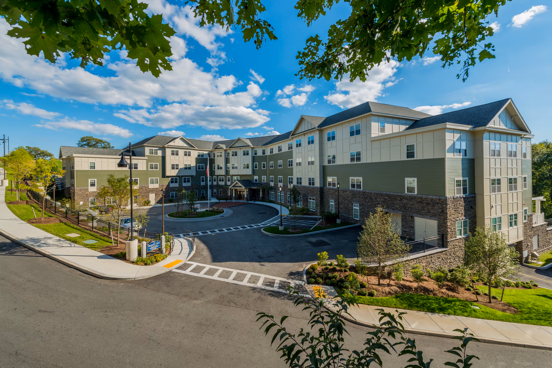 Front exterior of a multi-story senior living building with a circular driveway, landscaped grounds, and a bright blue sky.