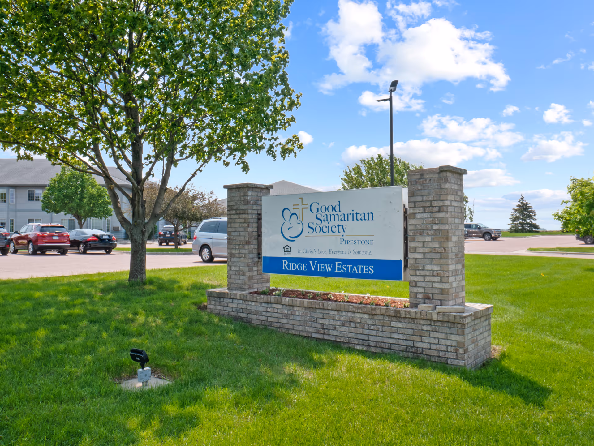 Outdoor view of the Good Samaritan Society - Pipestone - Ridge View Estates sign on a brick base surrounded by green grass and trees, with a parking lot and building in the background under a partly cloudy blue sky.