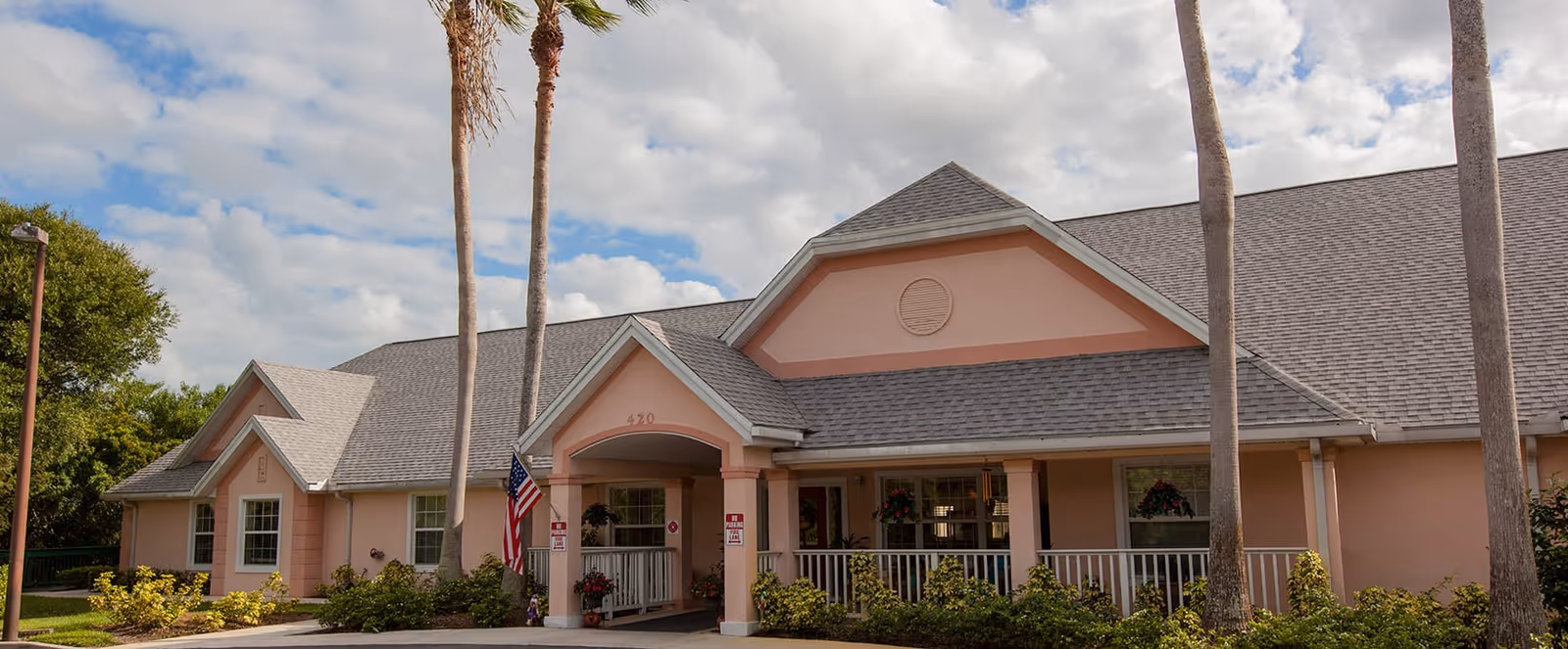 Exterior view of a single-story building with a gray shingled roof and peach-colored walls, featuring a covered entrance with white railings and columns, surrounded by palm trees and greenery under a partly cloudy sky.