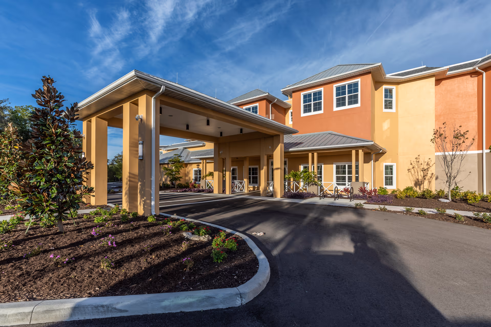 Exterior view of a senior living facility building with a covered entrance driveway, landscaped garden beds with small plants and flowers, and a clear blue sky above.