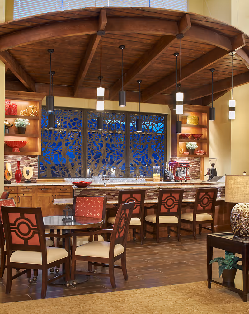 Interior view of a senior living facility dining area with a wooden ceiling and pendant lights. There is a bar counter with six chairs and a round table with four chairs in the foreground. The back wall features decorative shelving with red and gold accents and a coffee machine. A decorative window panel with a tree branch design is visible behind the bar.