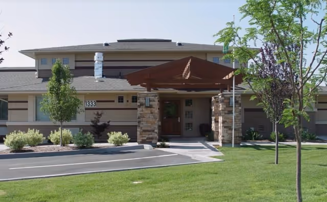 Front entrance of an assisted living building with a covered wooden porte-cochere, stone pillars, trees, and a manicured lawn.