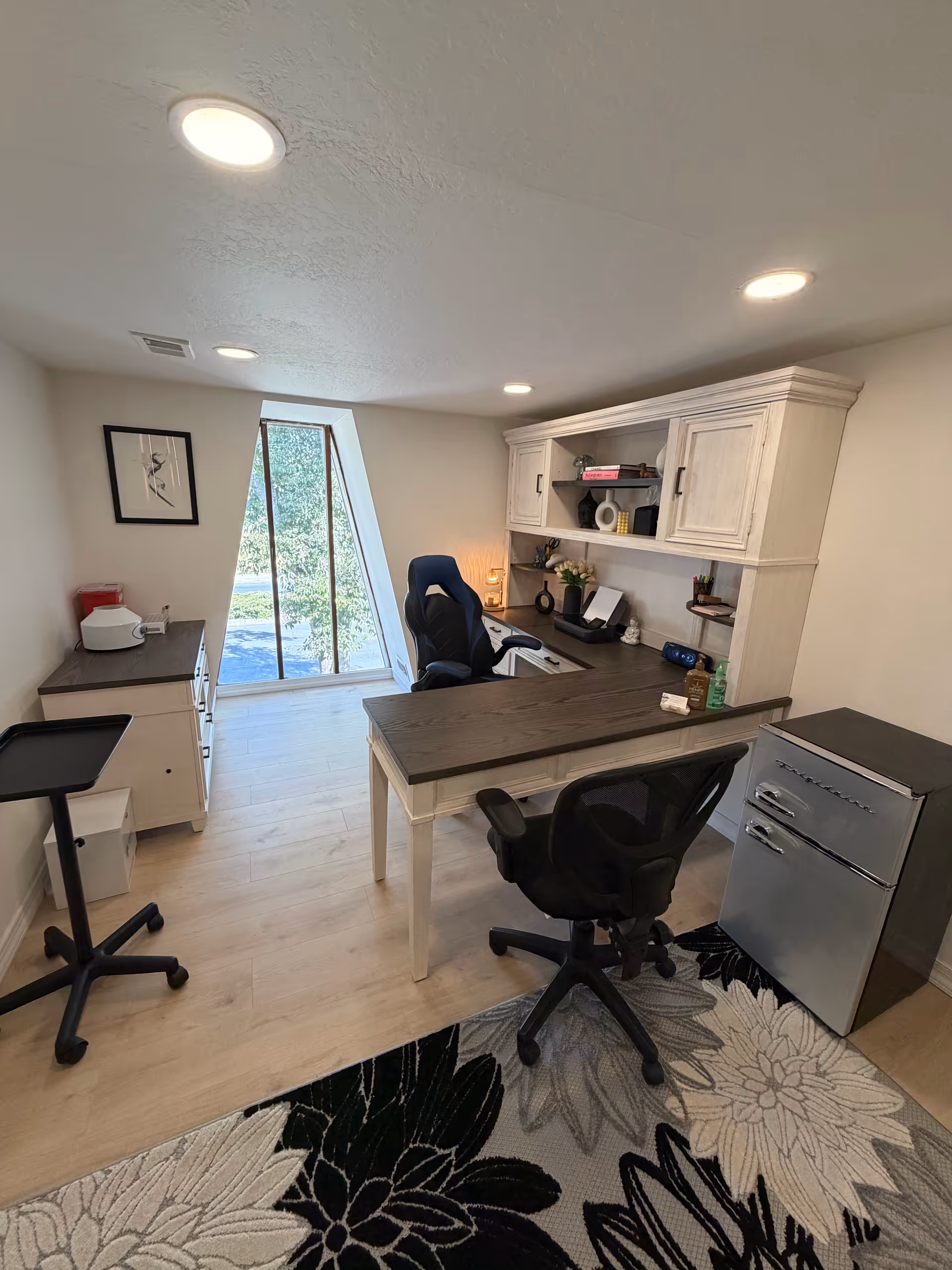 Bright office space with desk, chair, shelving, and a small fridge in a senior facility.