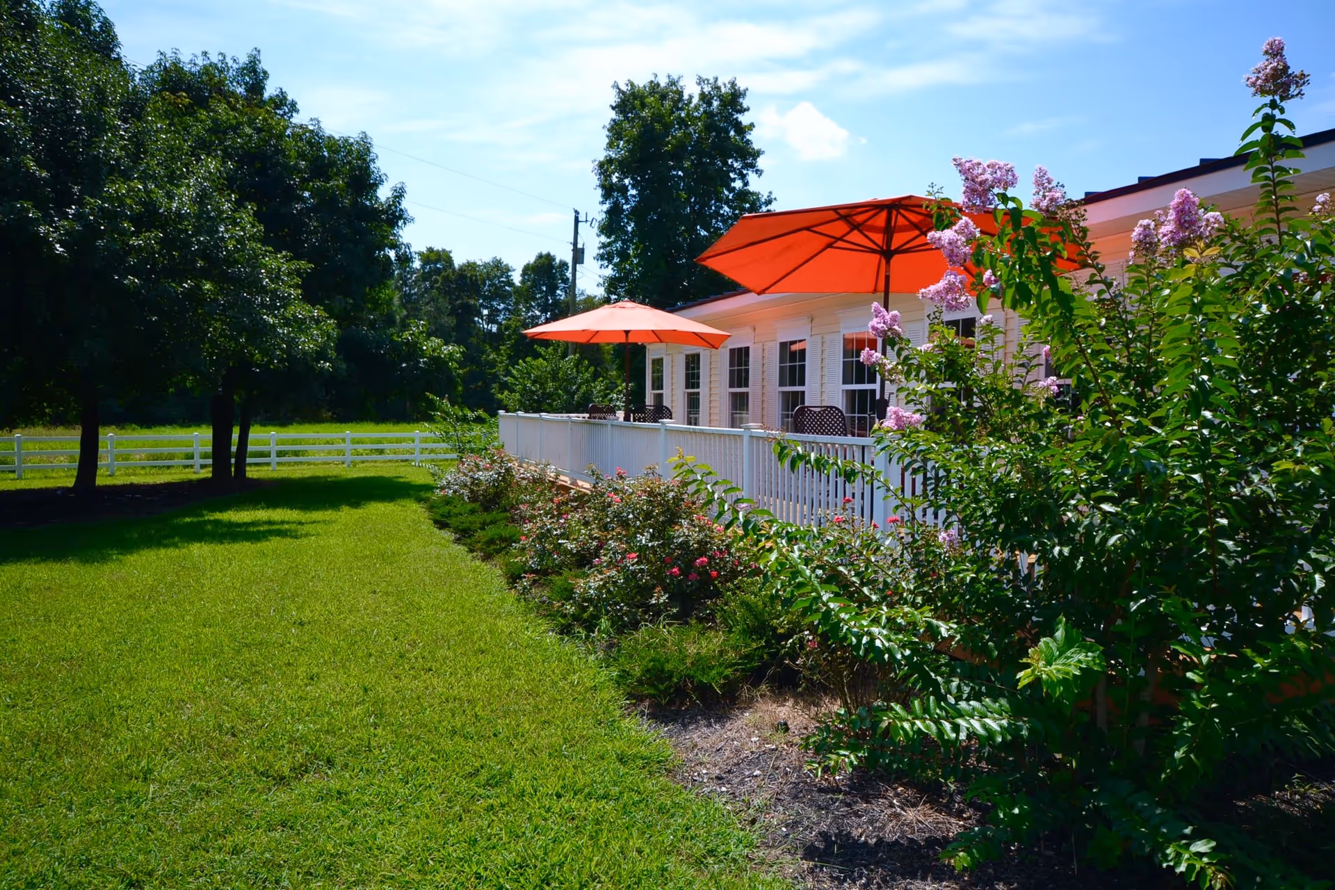 White single-story building with a porch, red patio umbrellas, flowering bushes and a manicured lawn.