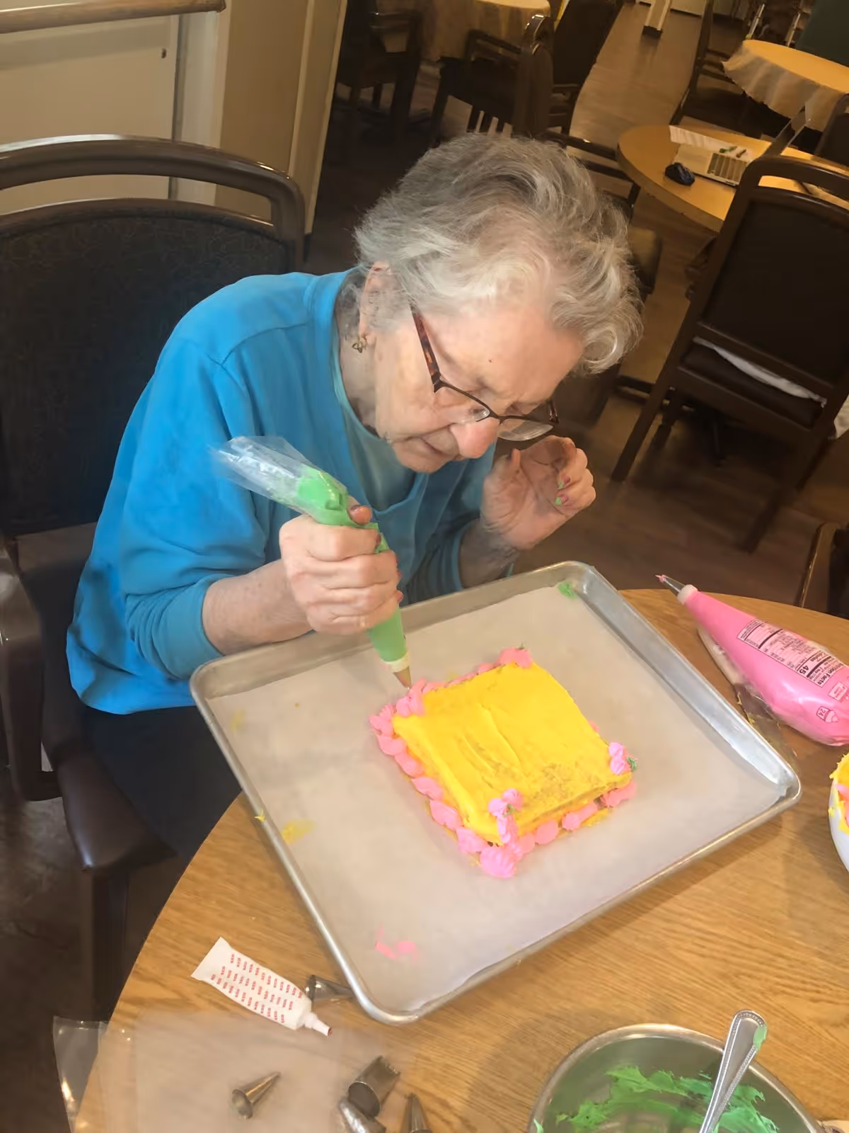 An elderly woman wearing glasses and a blue sweater is decorating a small square cake with yellow frosting and pink icing on a baking tray. She is sitting at a wooden table with various cake decorating supplies including piping bags and metal tips.