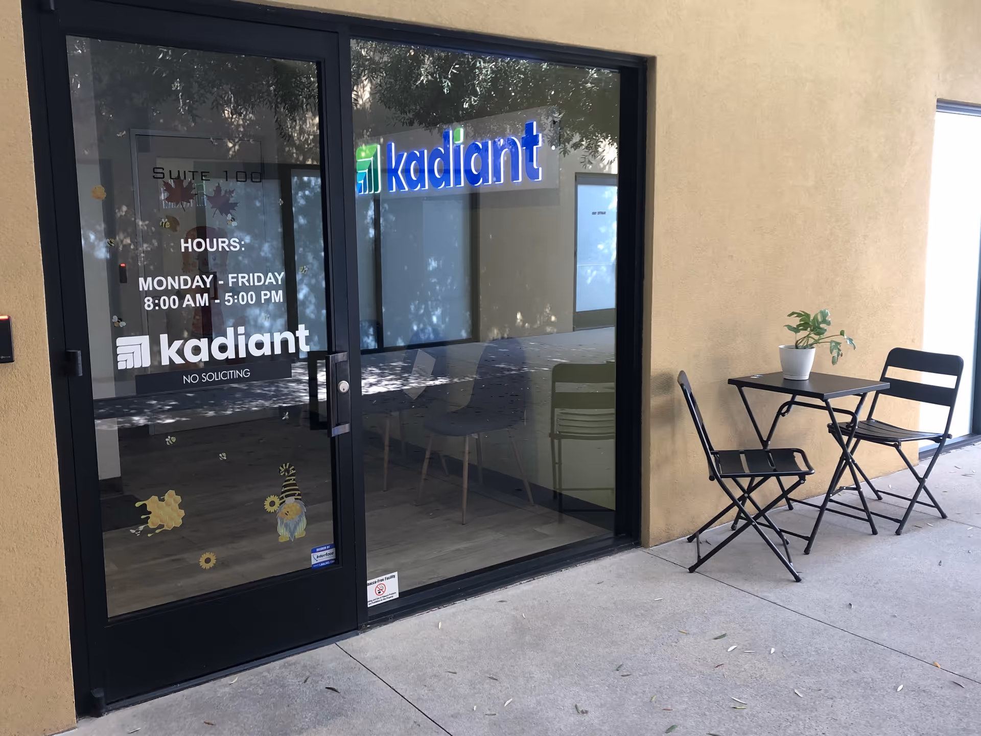 Exterior view of a Kadiant facility entrance with glass doors and a small outdoor table.