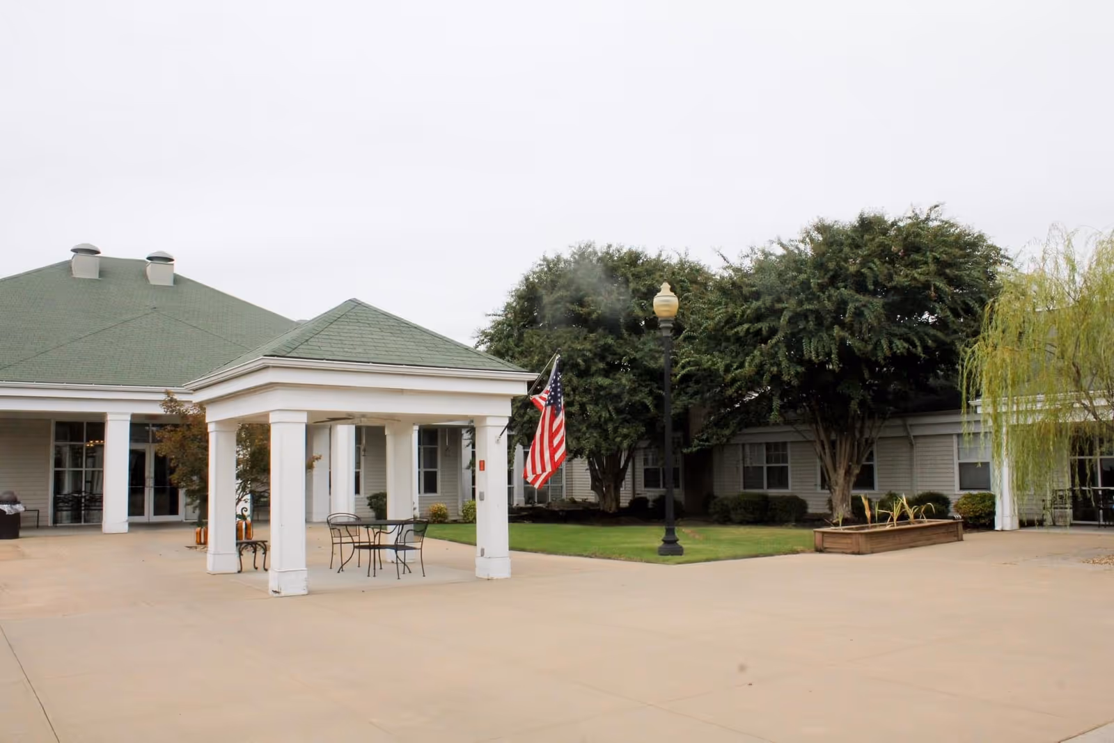 Front entrance of a senior living facility with a covered portico, American flag, outdoor seating, and surrounding trees.