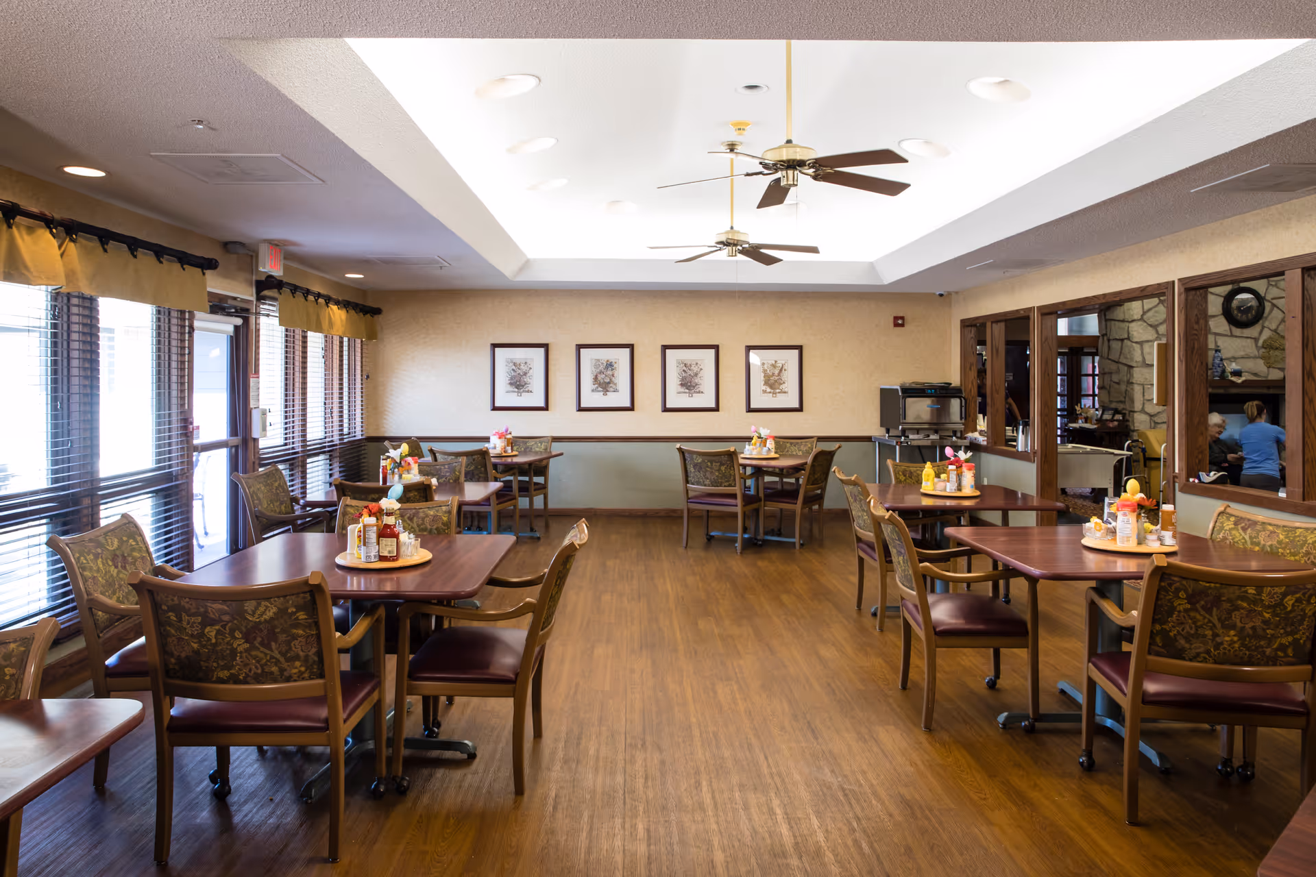 A well-lit dining room with several wooden tables and chairs arranged neatly. Each table has condiments and napkins placed on a round tray. Large windows with blinds and valances line the left side, allowing natural light to fill the room. The ceiling features recessed lighting and two ceiling fans. On the far wall, there are four framed pictures. A serving area with a coffee machine is visible on the right side, and a stone fireplace can be seen through a windowed partition.