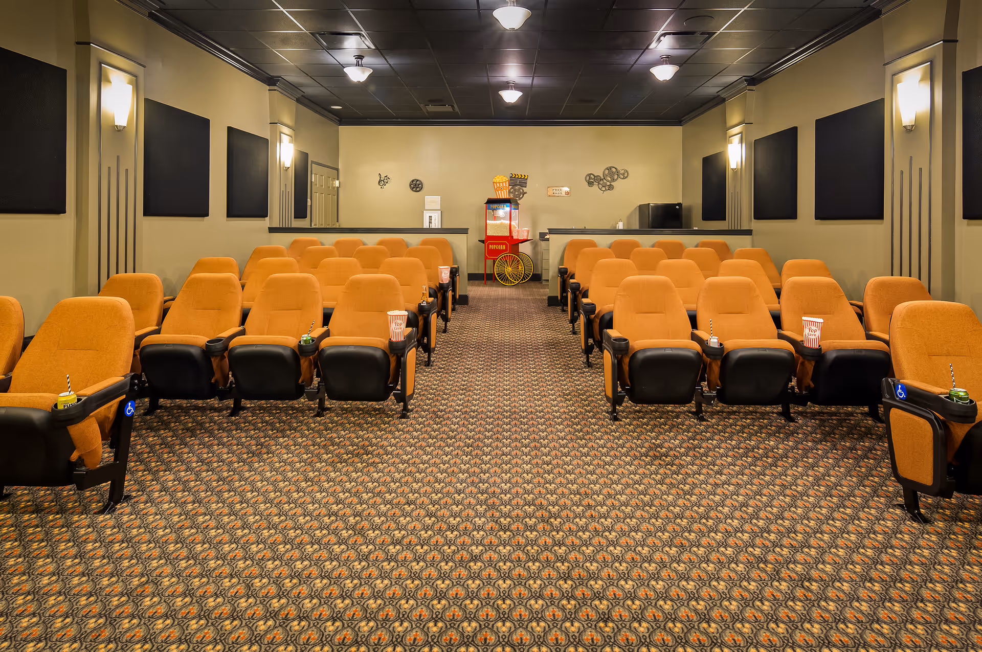 Interior view of a small movie theater room with rows of orange cushioned seats, each equipped with cup holders containing drinks and popcorn. At the back of the room, there is a popcorn machine and a small counter with a refrigerator. The walls are decorated with movie-themed wall art and soundproof panels.