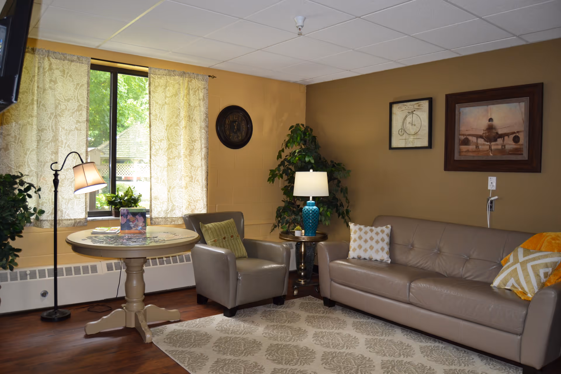 A cozy living room area with a beige leather sofa adorned with patterned pillows, a matching armchair with a green cushion, a round wooden table with a lamp and a small plant on it, and a floor lamp next to a window with sheer patterned curtains. The walls are painted brown and decorated with a clock and two framed pictures. A patterned rug covers part of the wooden floor.