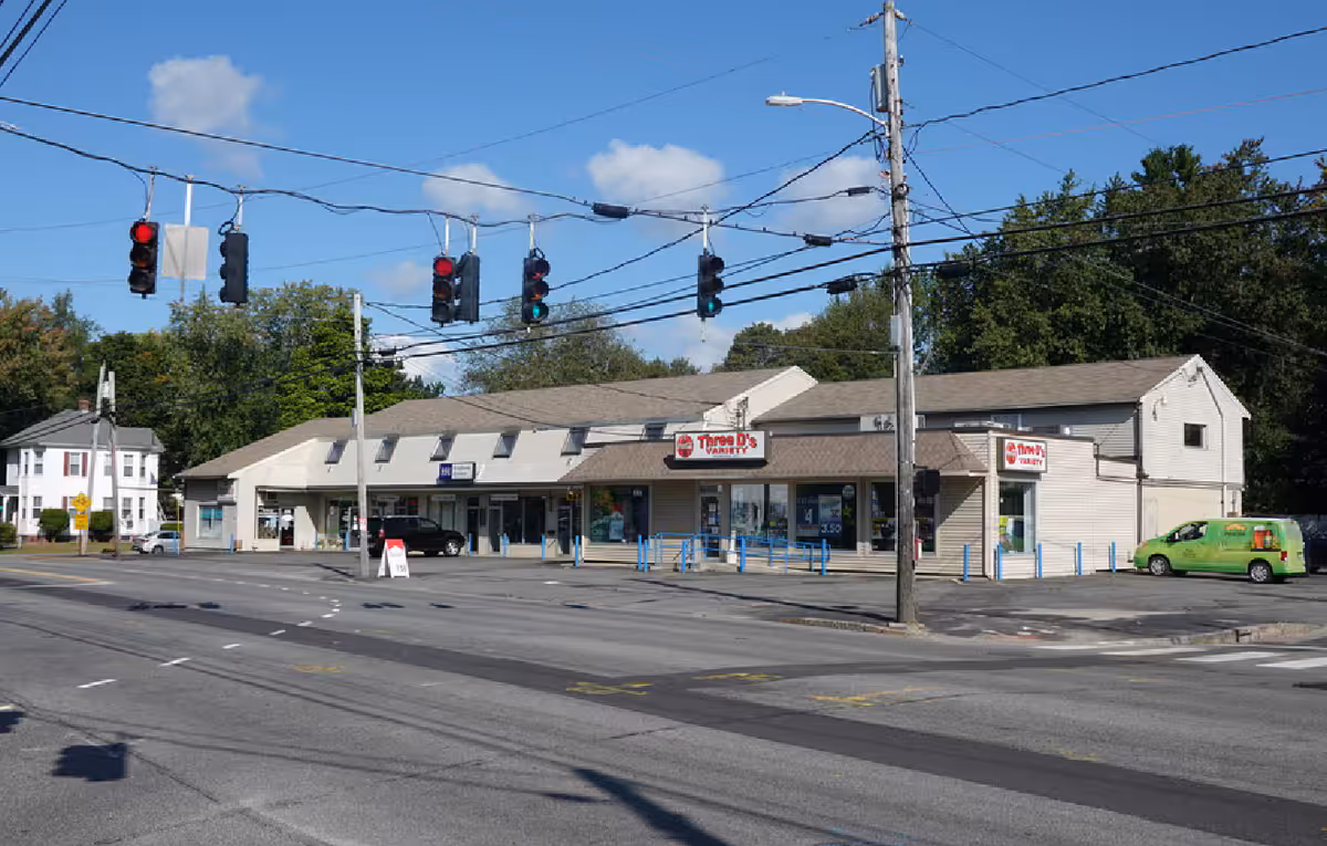 Exterior view of a strip mall with storefronts, traffic lights, and a green van.