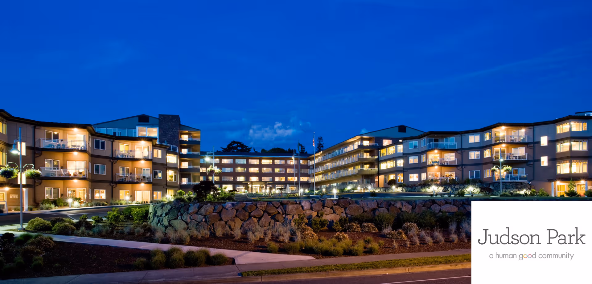 Exterior of a multi-story, well-lit senior living building at dusk with landscaped rockery and pathways, and a Judson Park logo in the corner.