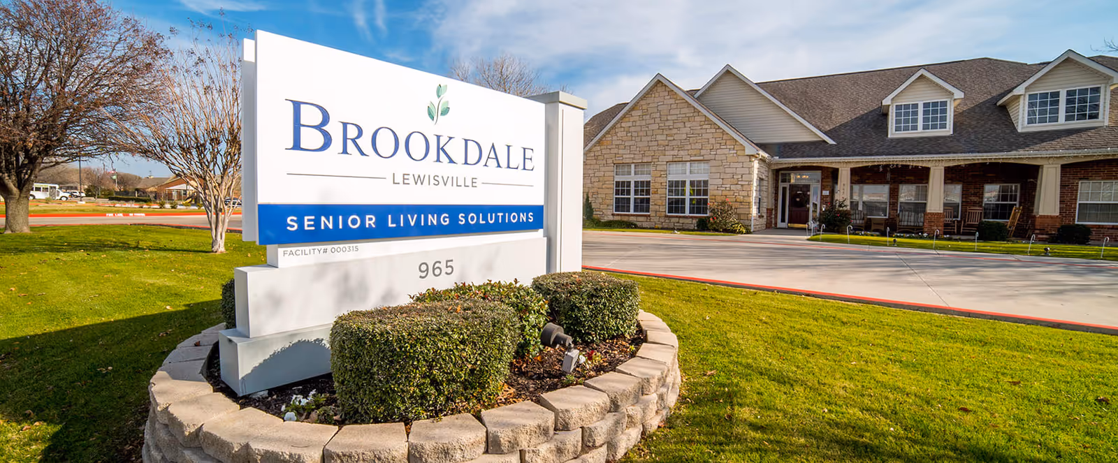 Front exterior of the Brookdale Lewisville senior living facility with a large sign reading 'Brookdale Lewisville Senior Living Solutions' on a landscaped lawn.