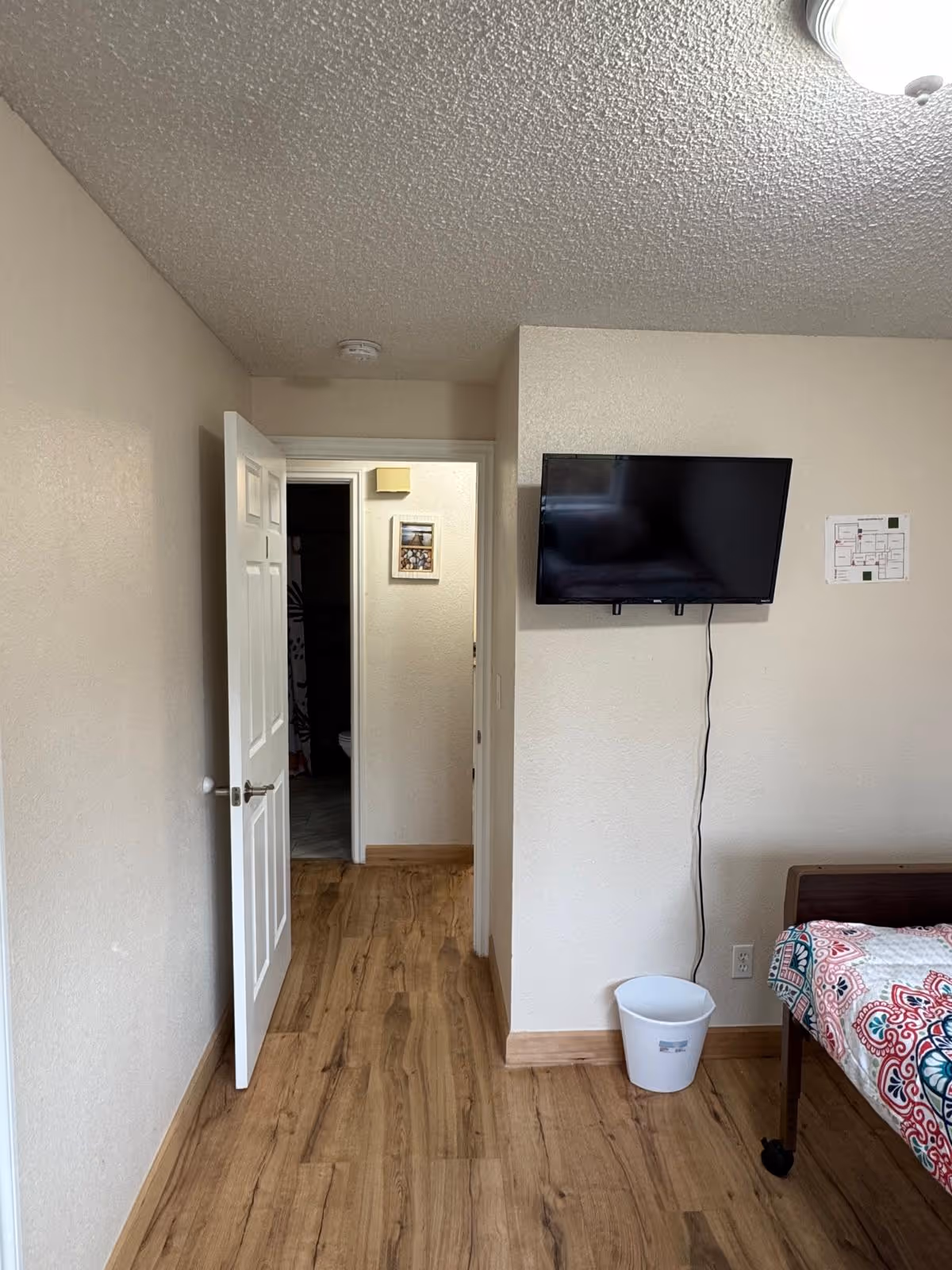 Interior view of a bedroom with a mounted TV, doorway, bed, and wooden floor.