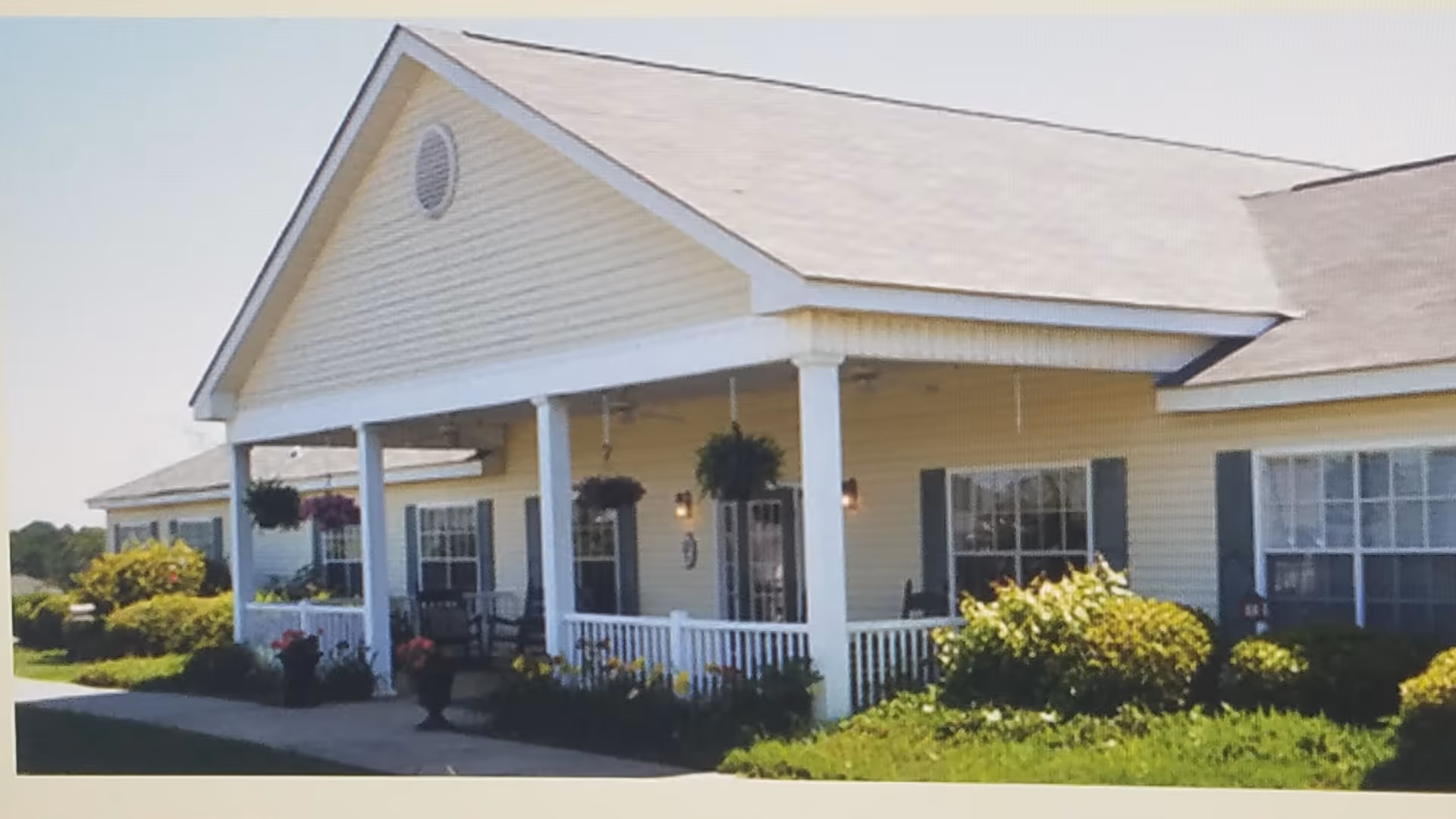 Exterior view of a single-story senior living facility building with a covered porch, white columns, hanging flower baskets, and surrounding greenery and flowers.