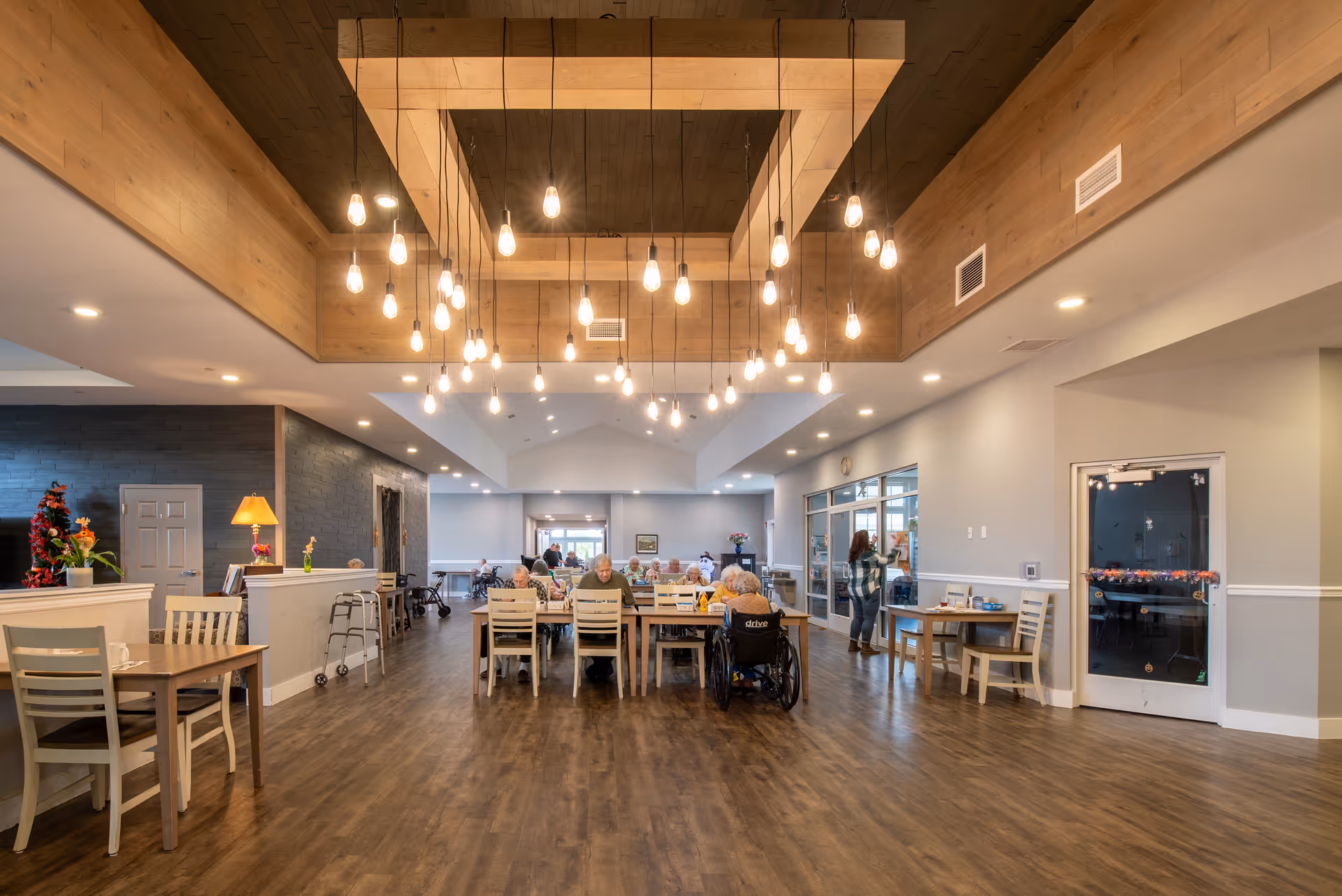 A spacious dining room in a senior living facility with wooden floors and a modern wooden ceiling feature with hanging light bulbs. Several elderly residents are seated at tables, some in wheelchairs, engaging in activities or dining. A woman is standing near a glass door on the right side. The room is well-lit with recessed lighting and has a cozy, welcoming atmosphere.