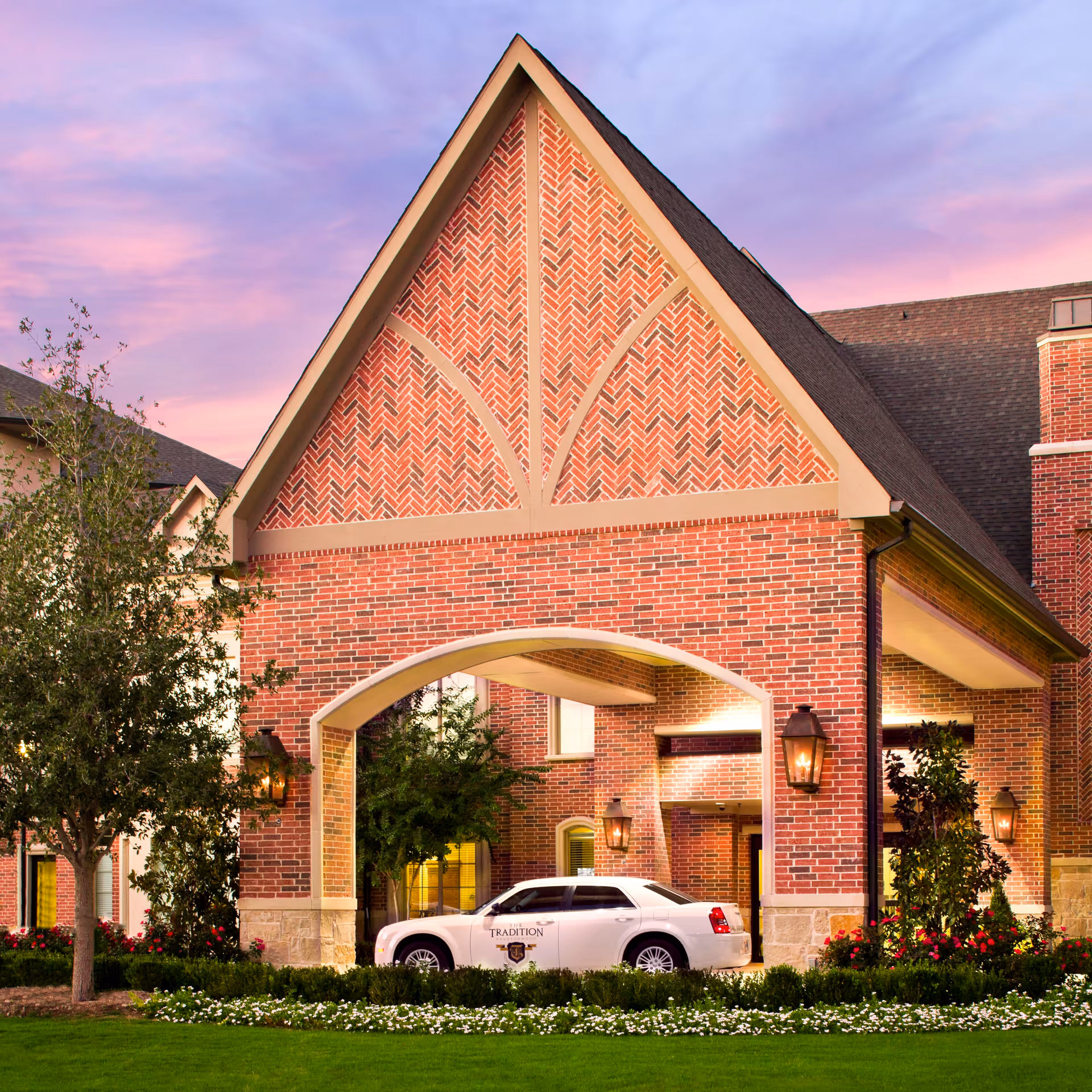 Exterior view of a senior living facility entrance with a large brick archway and a white car parked underneath. The building features red brick walls, decorative brick patterns, and outdoor lantern-style lights. There is a well-maintained lawn and landscaping with trees and flowers in front of the entrance, under a pink and purple sunset sky.