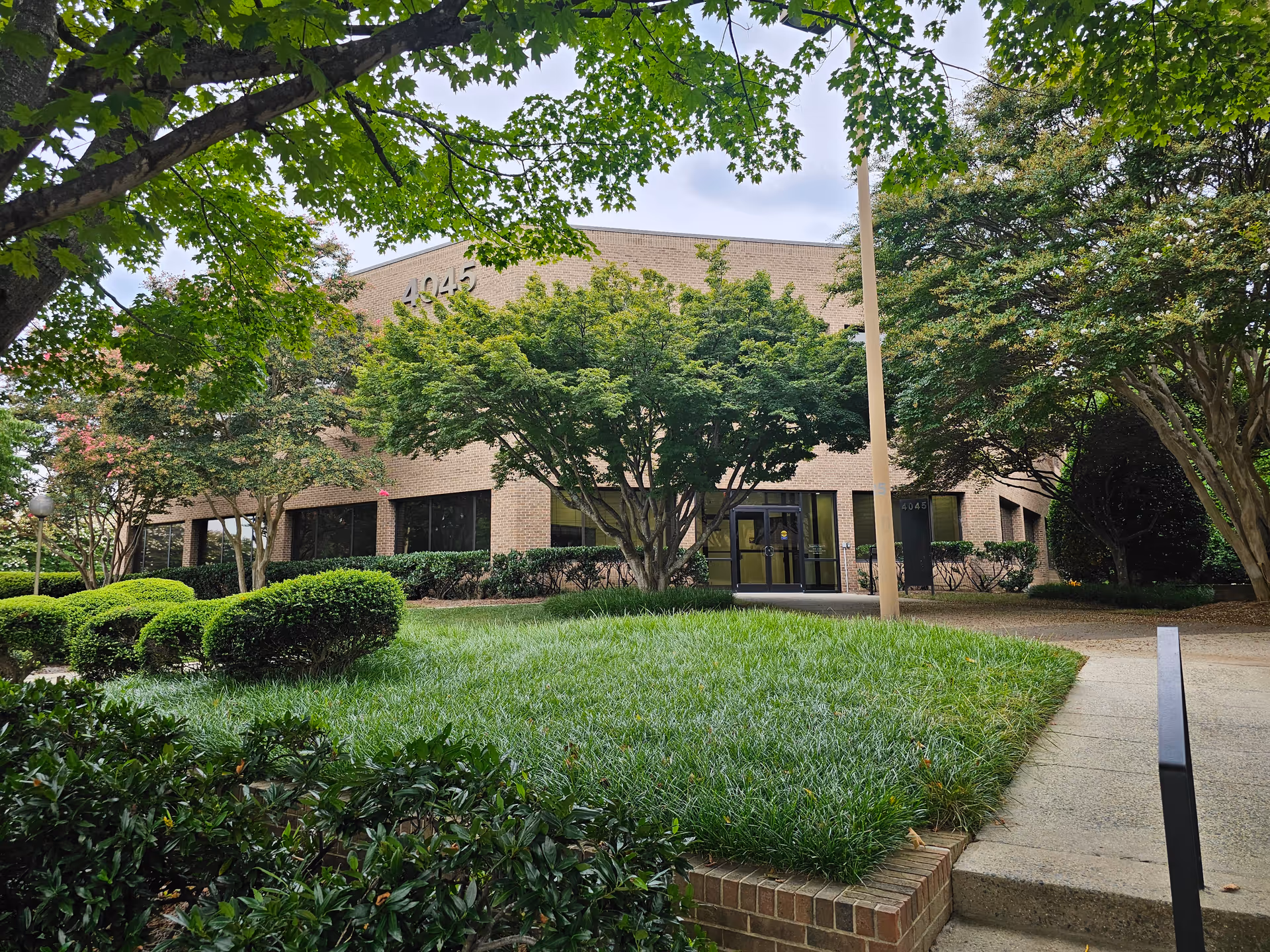 Brick building with glass entrance, surrounded by green shrubs and trees.