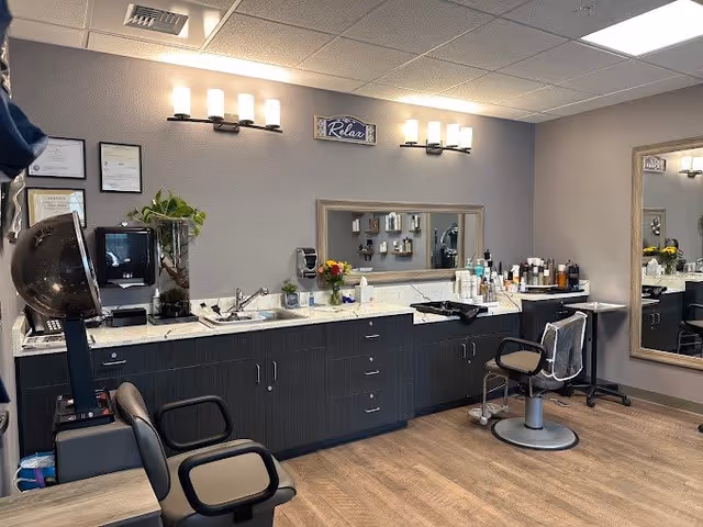 Interior view of a senior living facility salon area with two salon chairs, a hair dryer, a large mirror on the wall, cabinets with various hair care products, and soft lighting fixtures above. The walls are painted light gray and the floor has wood-like finish. A small sign that says 'Relax' is mounted above the mirror.