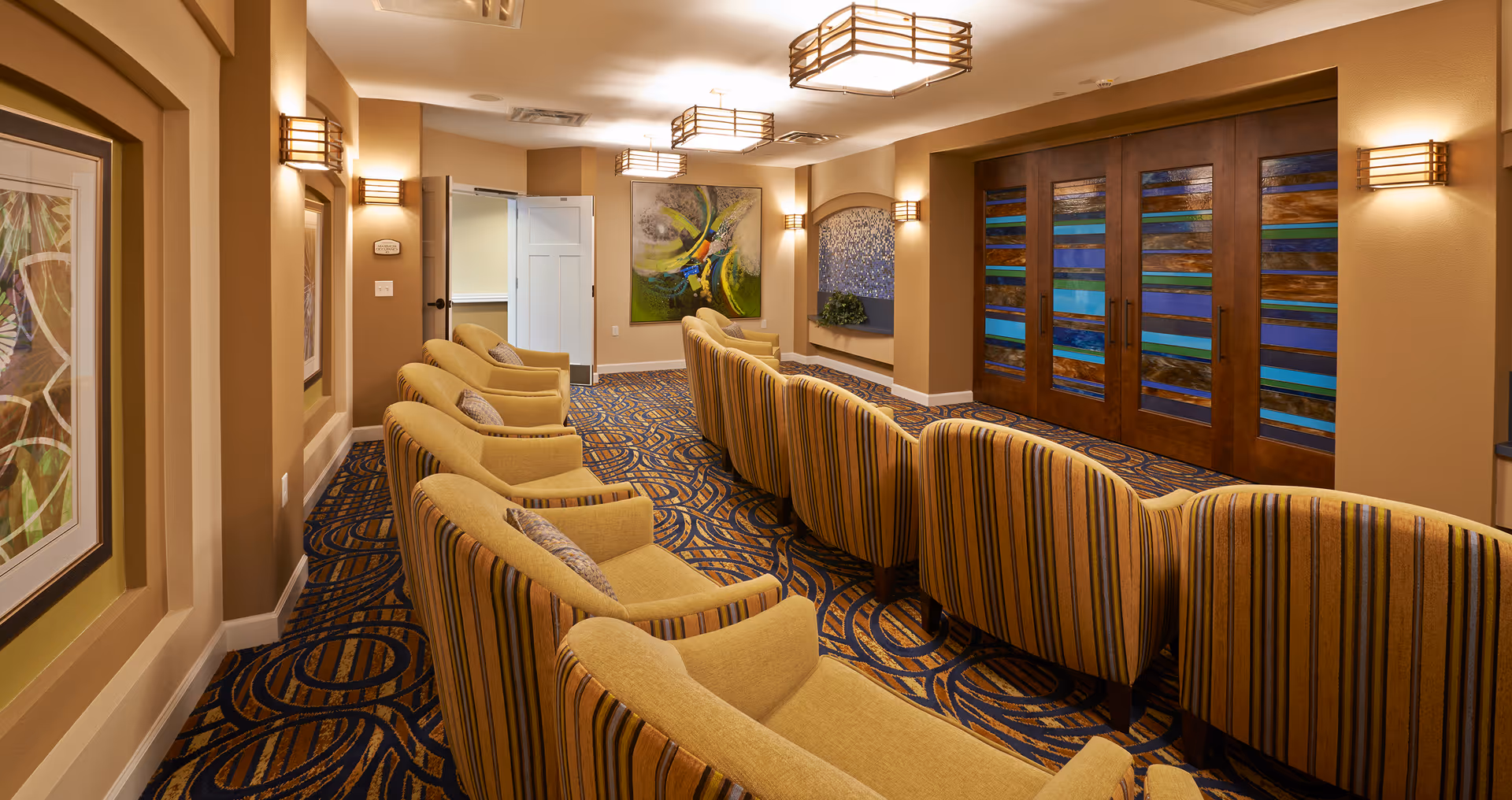 A cozy interior lounge area with two rows of striped yellow armchairs facing a wall with decorative wooden panels featuring blue and green glass inserts. The room has warm beige walls, patterned carpet with blue and brown swirls, framed artwork, and soft wall-mounted lighting fixtures. An open door leads to another room in the background.