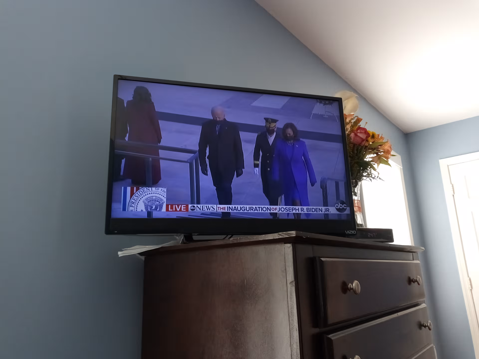 Television on a dresser with flowers against blue walls, displaying the Biden inauguration.
