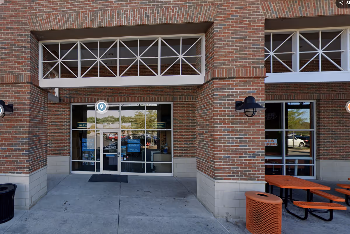 Brick storefront exterior with glass doors, windows, and orange outdoor tables near the entrance.