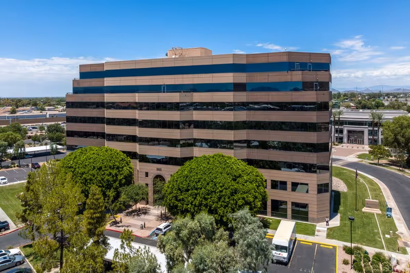 A multi-story tan and glass office building with trees and a parking lot in foreground.