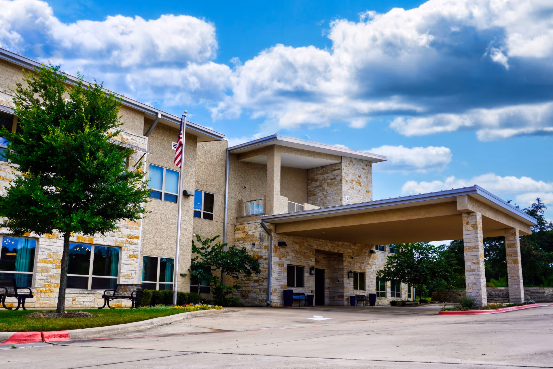 Exterior view of a senior living facility building with beige stone and stucco walls, large windows, a covered entrance supported by stone pillars, an American flag on a flagpole, green trees, and a partly cloudy blue sky.