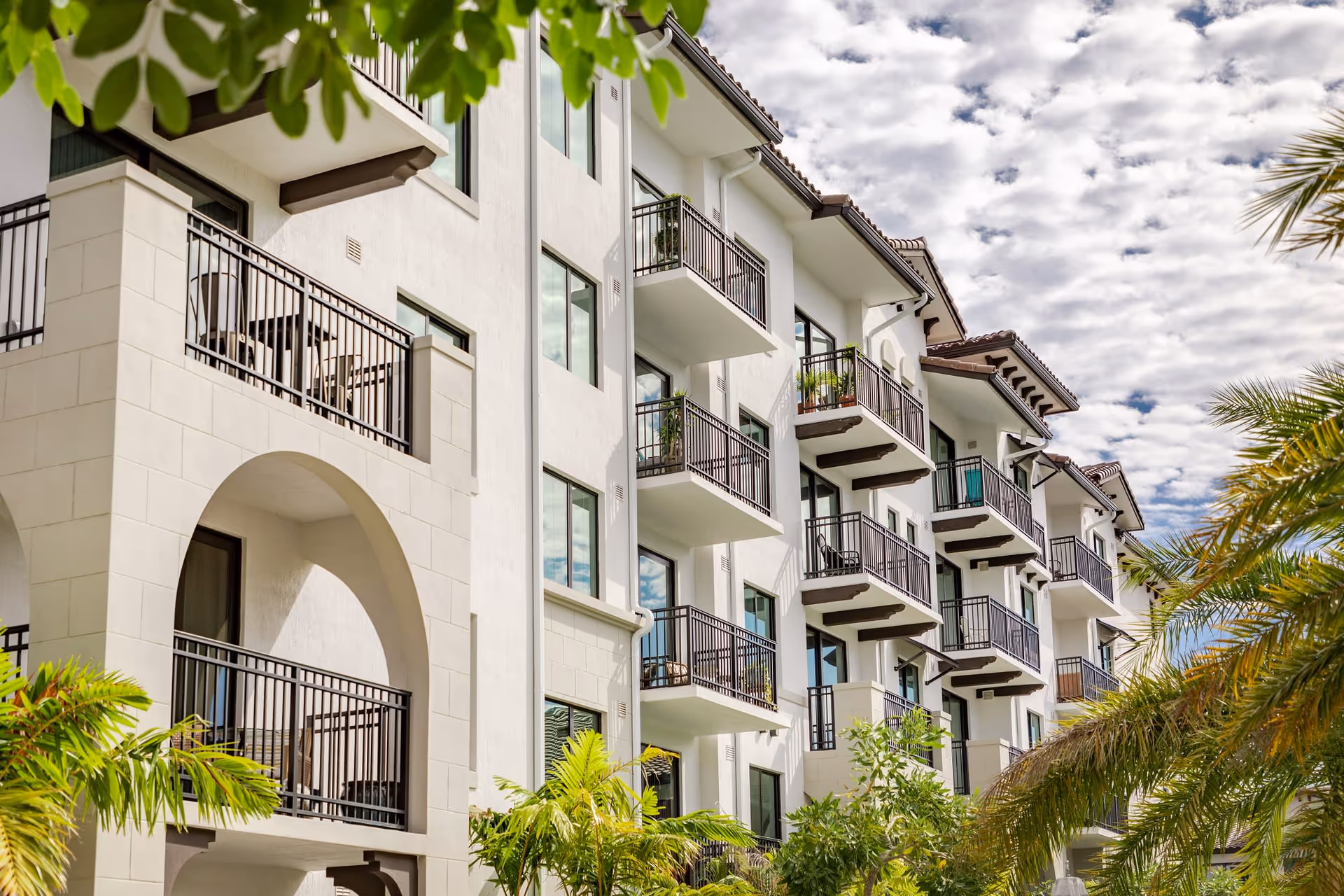 White Mediterranean-style multi-story residential building with balconies and palm trees under a partly cloudy sky.
