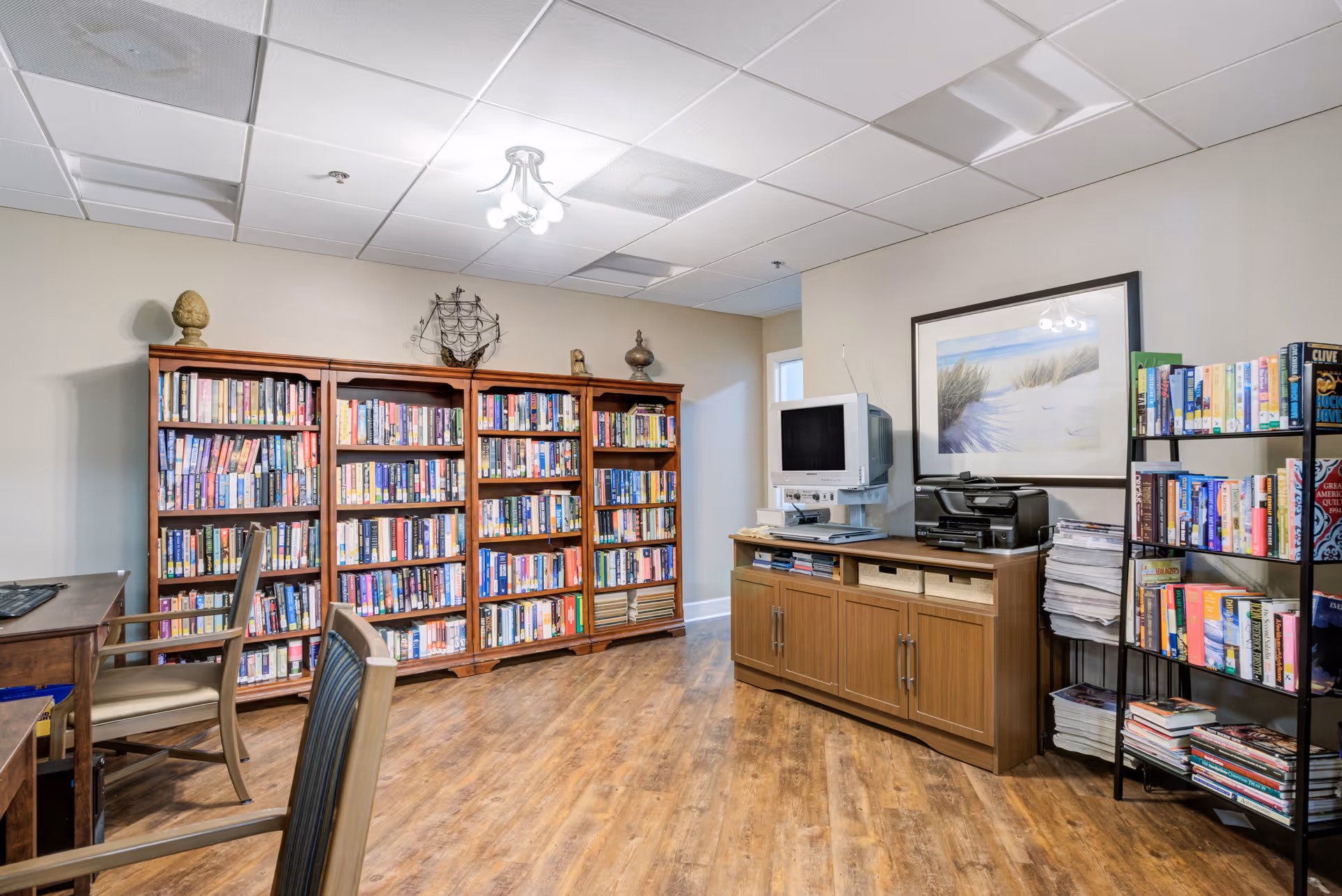 A well-lit senior living facility library with tall bookshelves, a computer desk, and magazines.