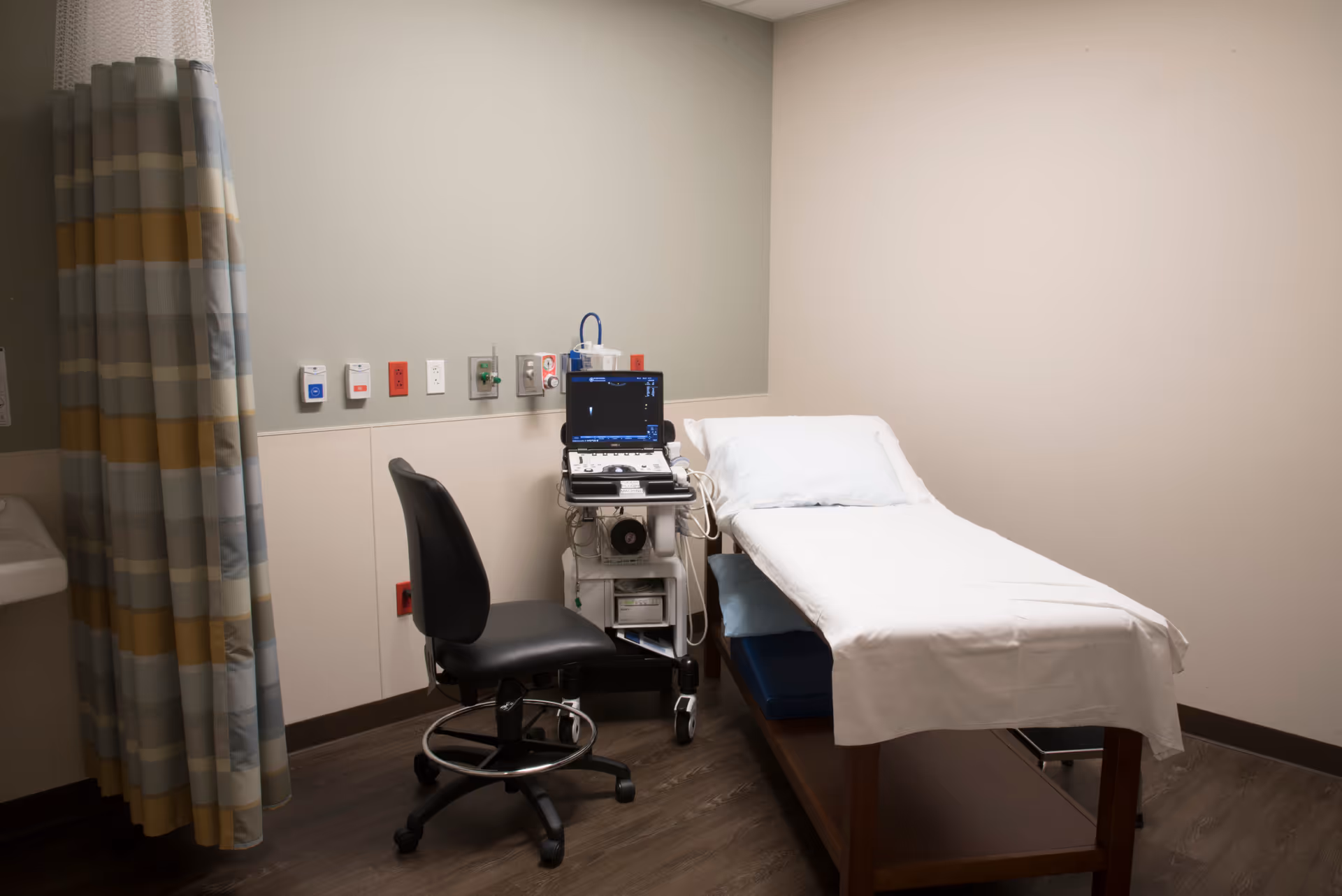 A clinical examination room featuring a bed, medical monitor on a cart, and an adjustable chair.