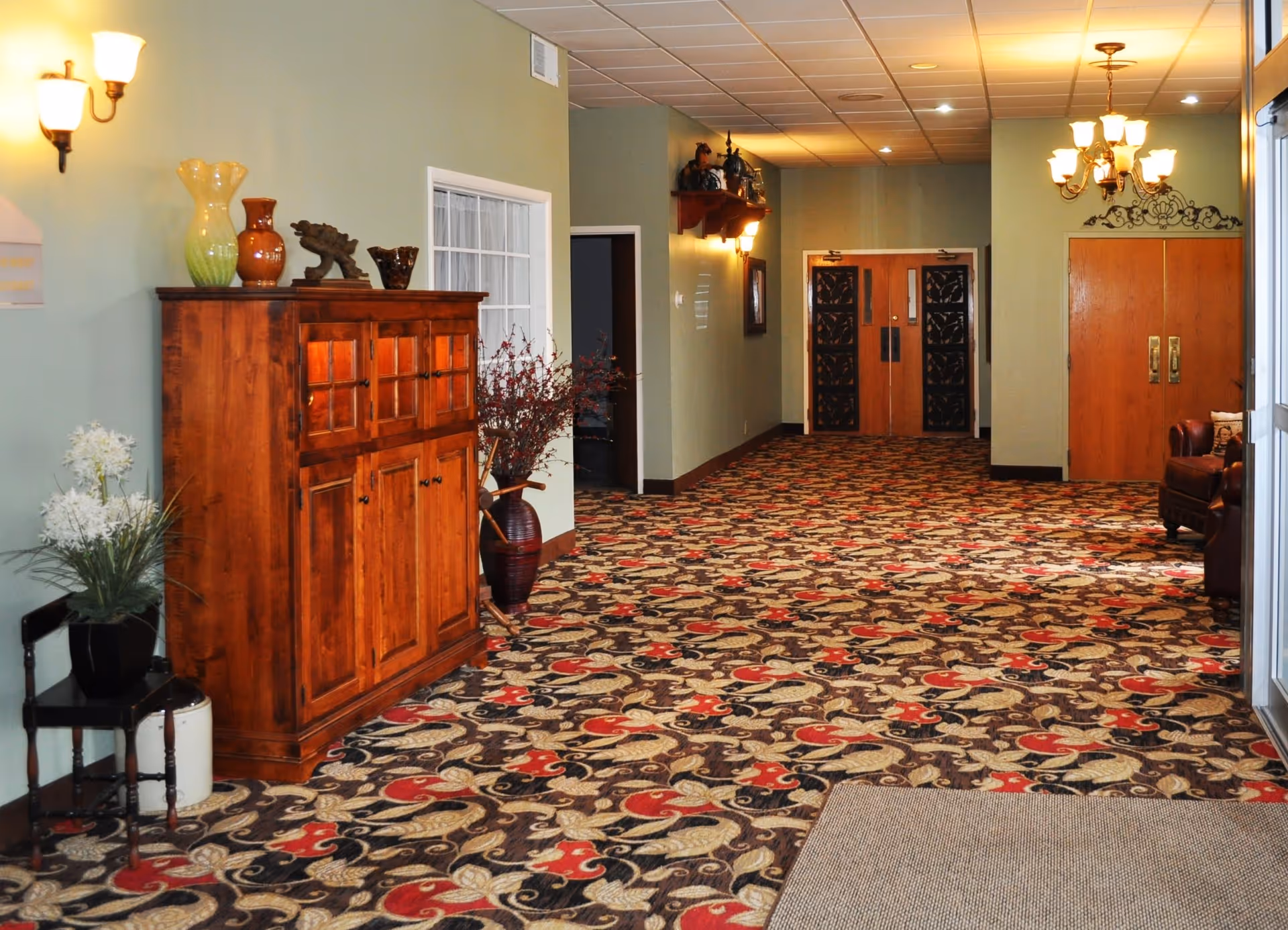 Interior hallway of a senior living facility with patterned carpet, light green walls, wooden cabinet with decorative vases, wall sconces, and double wooden doors at the end. There is a chandelier and some seating visible on the right side.