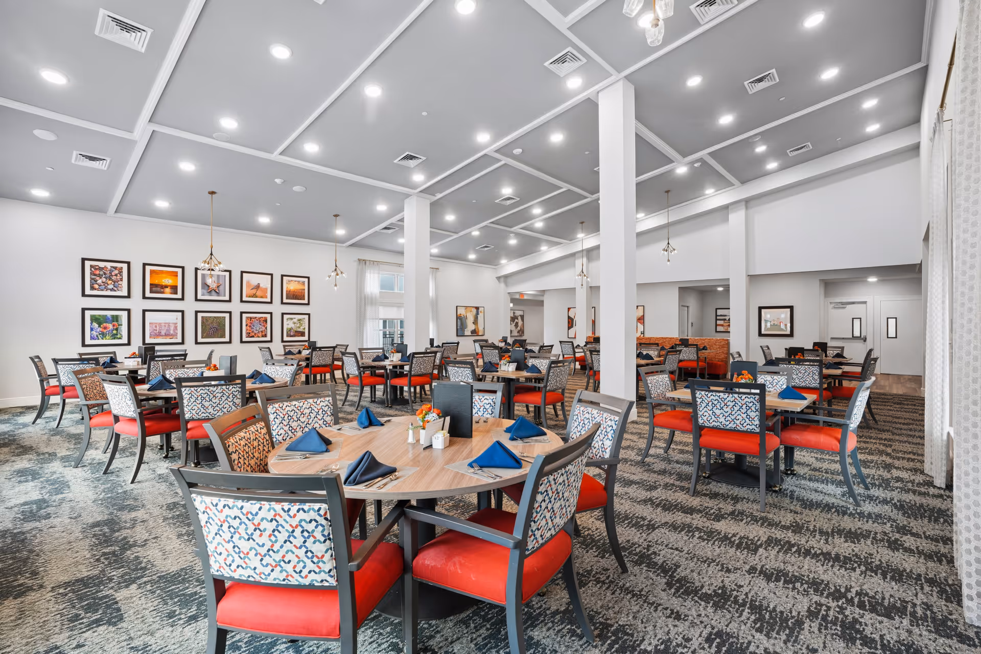 Bright dining hall with round tables, patterned chairs, napkins, and framed wall art.