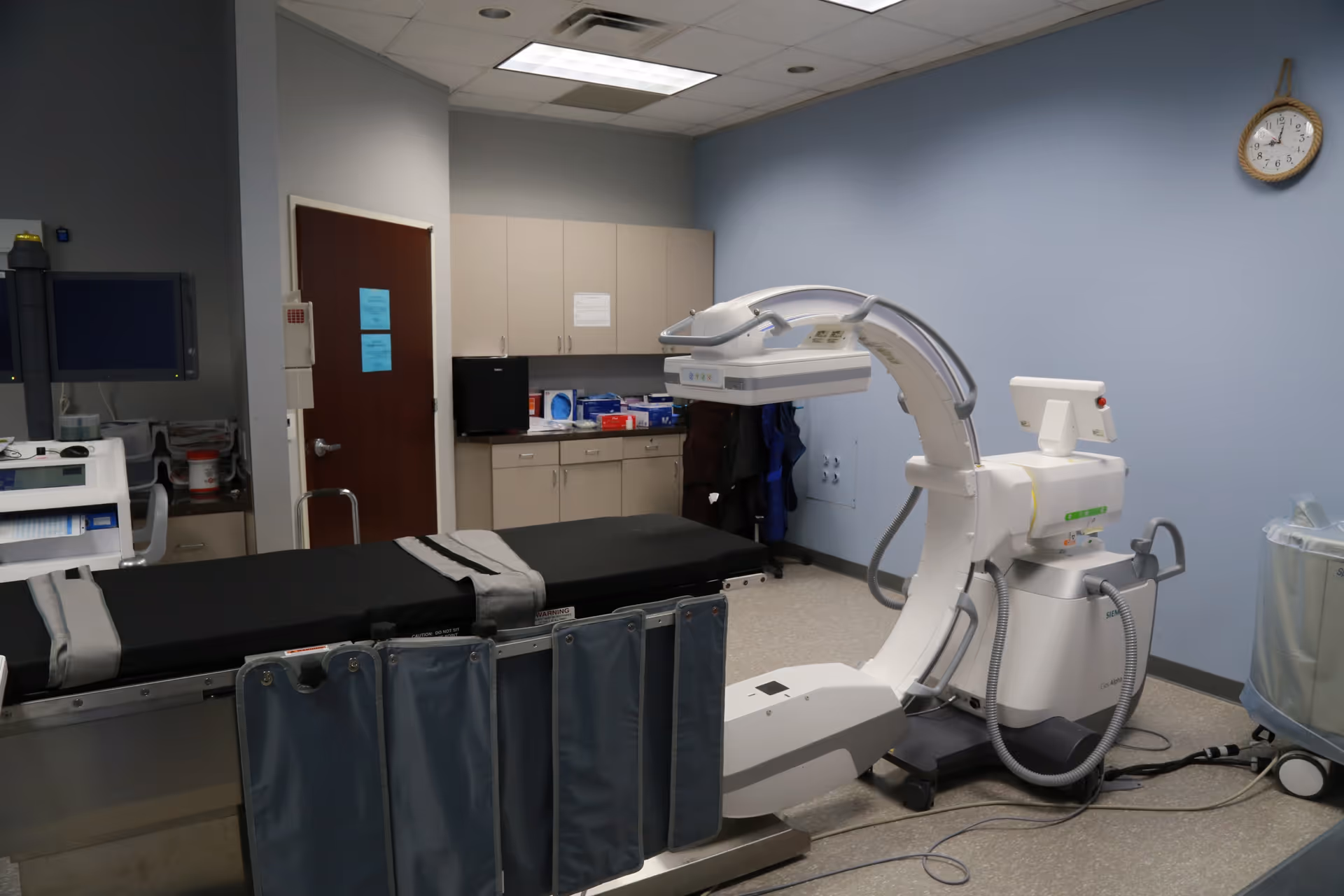 Medical imaging suite with a patient bed, angiography machine, cabinets, and clock on blue wall.
