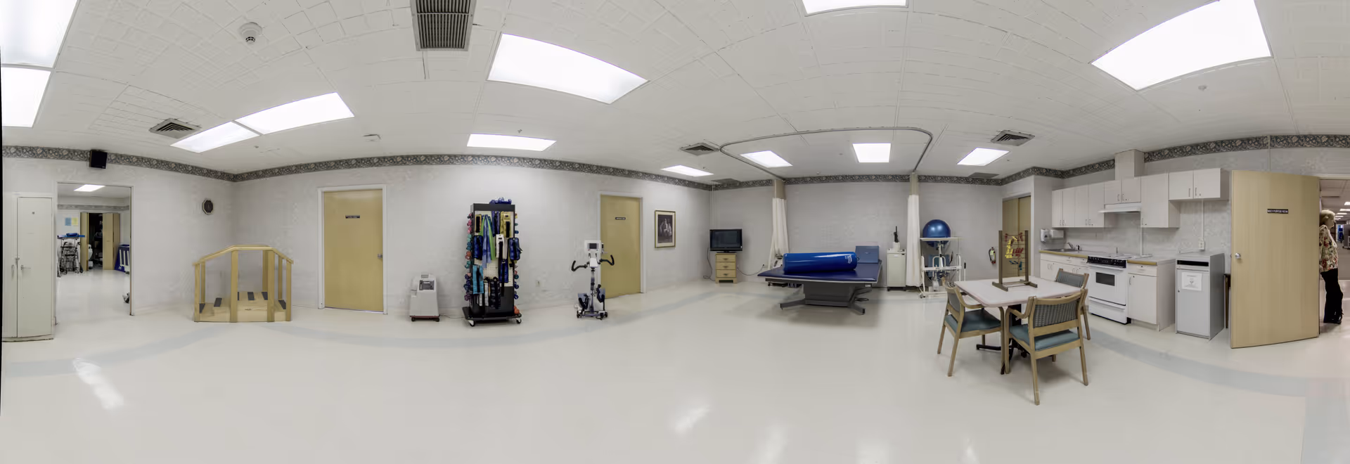 Wide interior view of a senior living facility with a therapy table, kitchenette, and dining area.