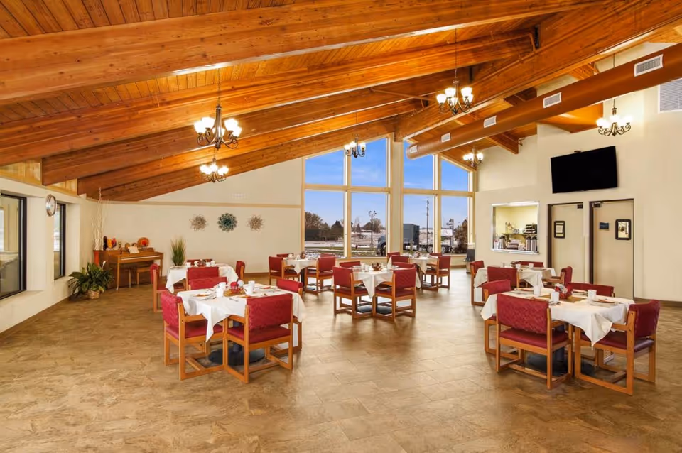 A spacious dining room with wooden vaulted ceilings and large windows letting in natural light. The room contains multiple tables covered with white tablecloths, each surrounded by red cushioned chairs. There are chandeliers hanging from the ceiling, a piano in the corner, and a television mounted on the wall near double doors.