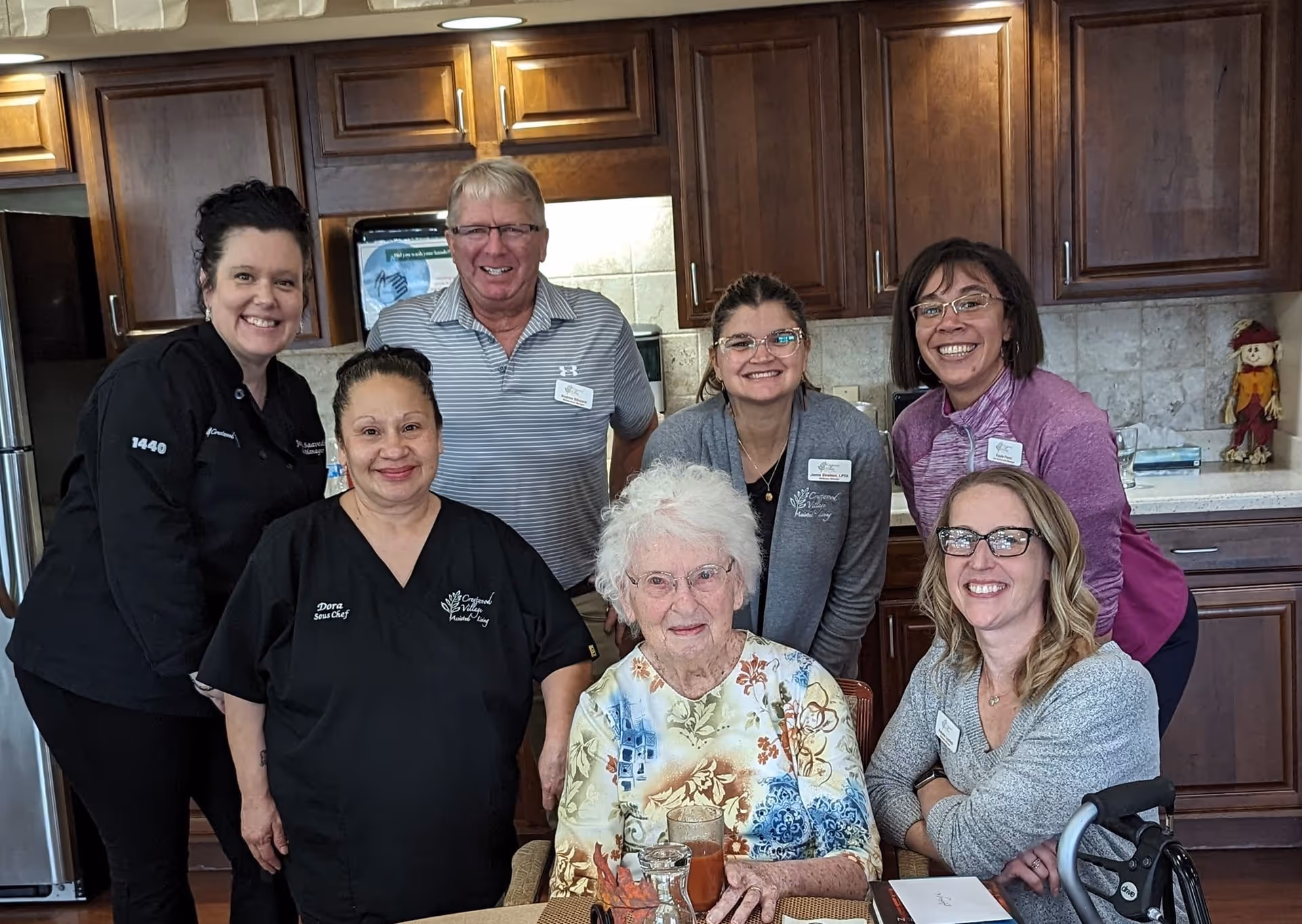 A group of seven people, including an elderly woman seated at a table and six staff members standing and sitting around her, smiling in a kitchen area with wooden cabinets and a refrigerator in the background.