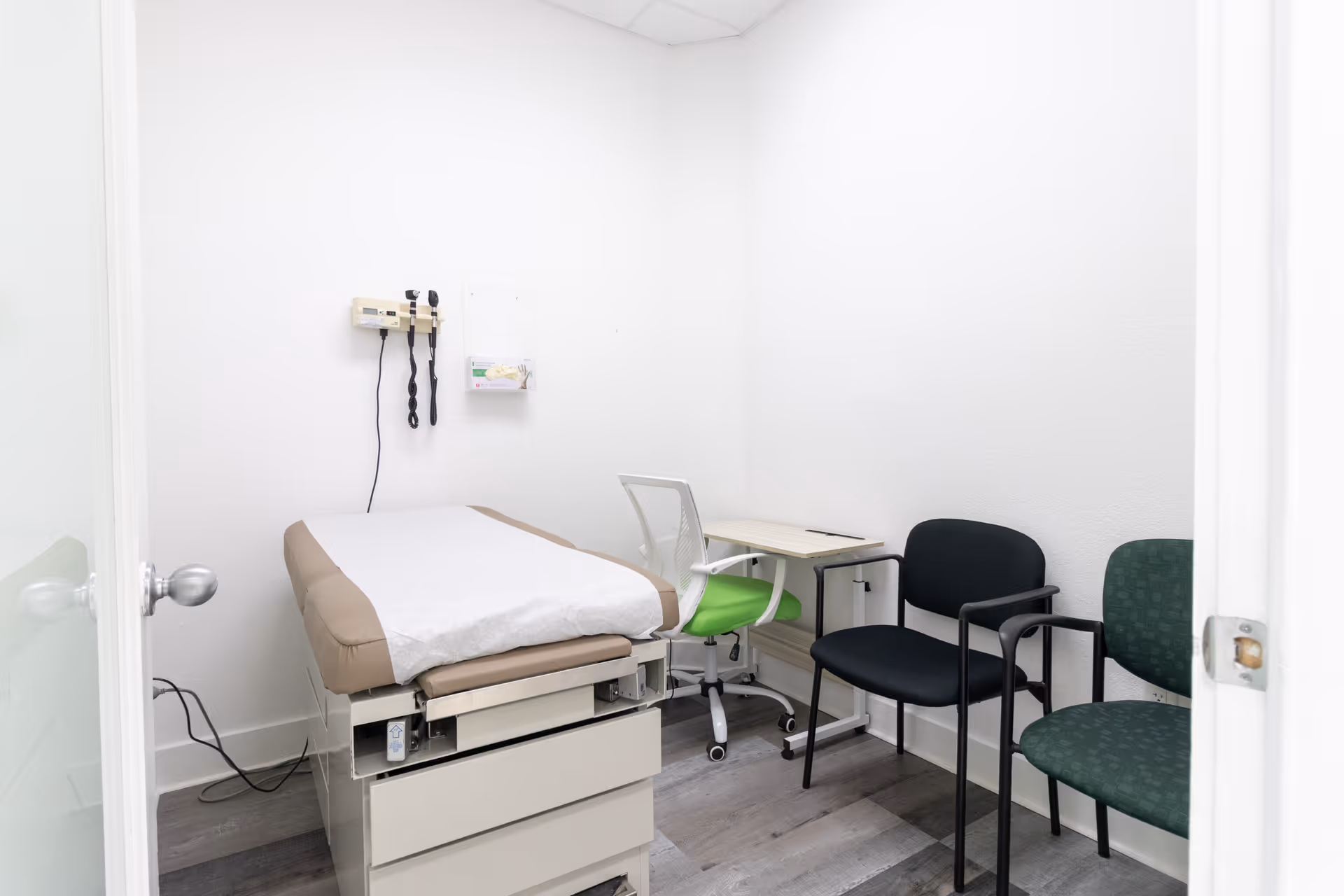 Clinical examination room with an exam bed, desk, and several chairs.