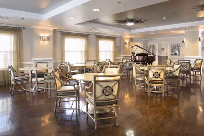 Well-lit senior living dining room with round tables, chairs, and a grand piano.