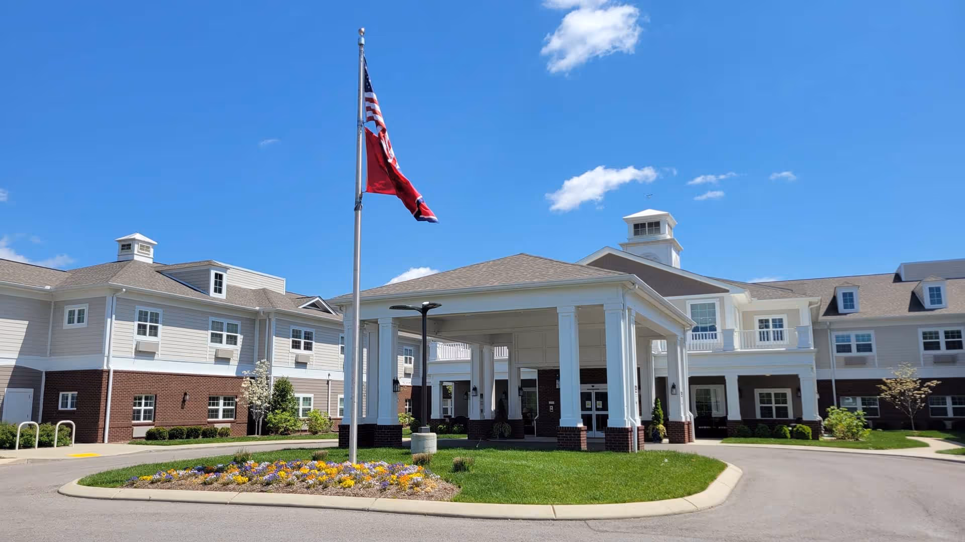 Exterior view of The Goldton at Spring Hill senior living facility on a clear sunny day, featuring a large covered entrance with white columns, a flagpole with an American flag and another flag, landscaped flower beds, and a multi-story building with beige siding and brick accents.