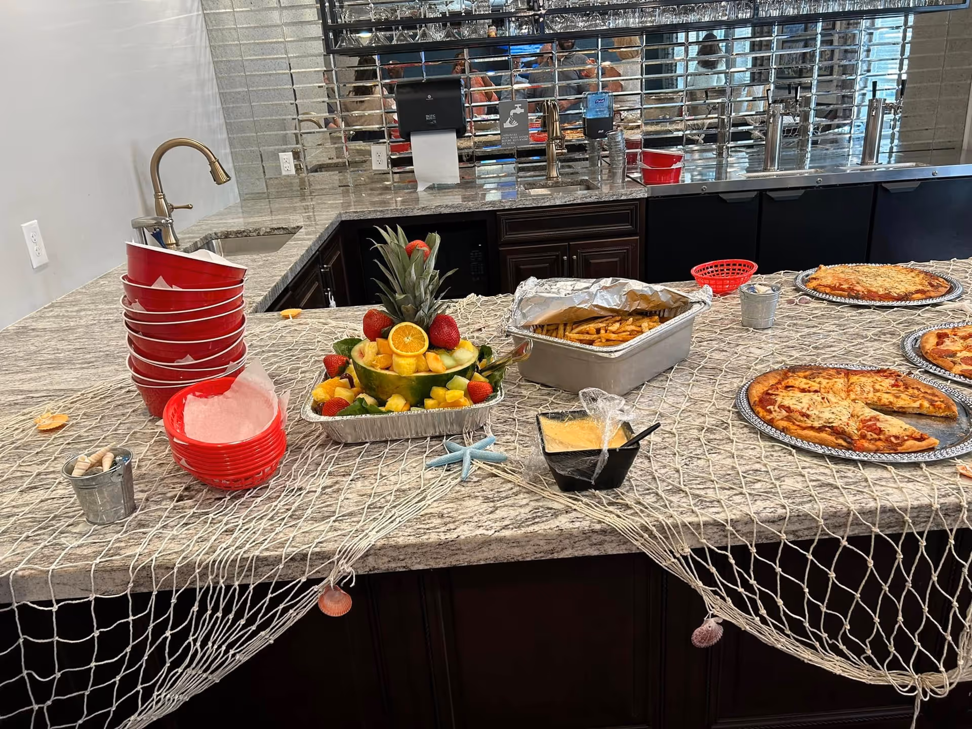 A buffet-like kitchen island with fruit, pizzas, cups, and food trays.