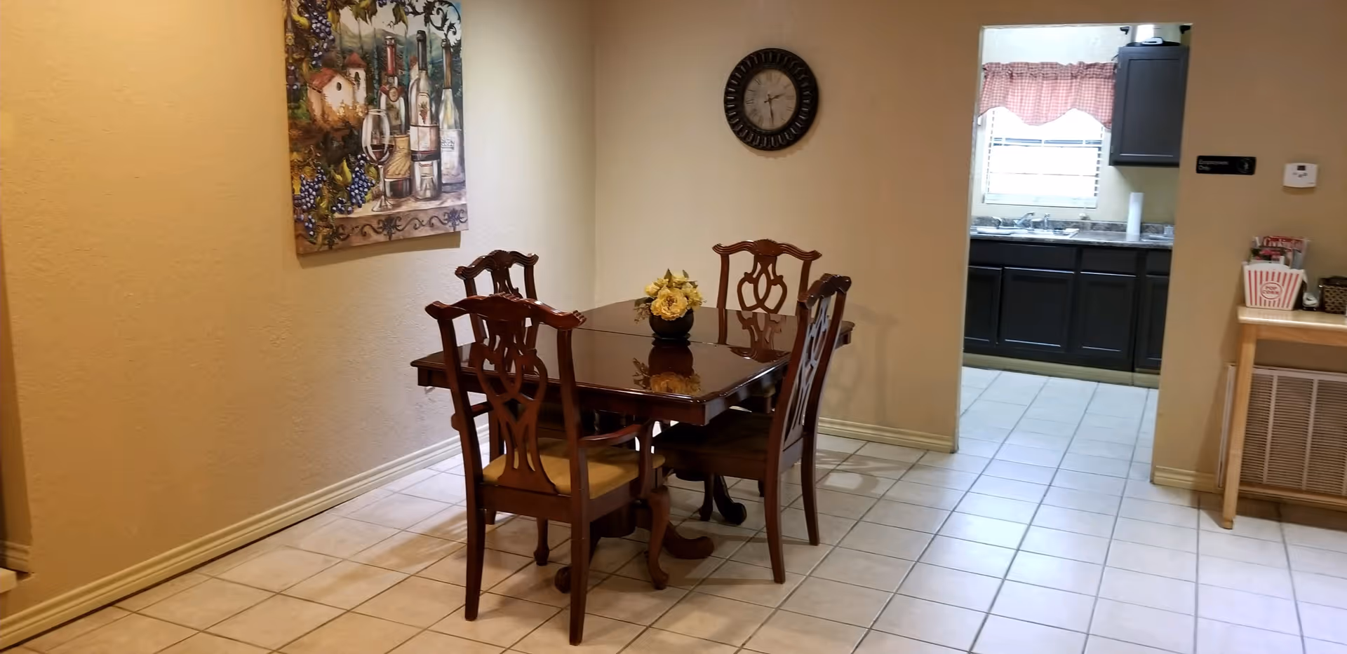 Small dining area with a wooden table and four chairs on a tiled floor, wall clock and artwork, and a view into the kitchen.