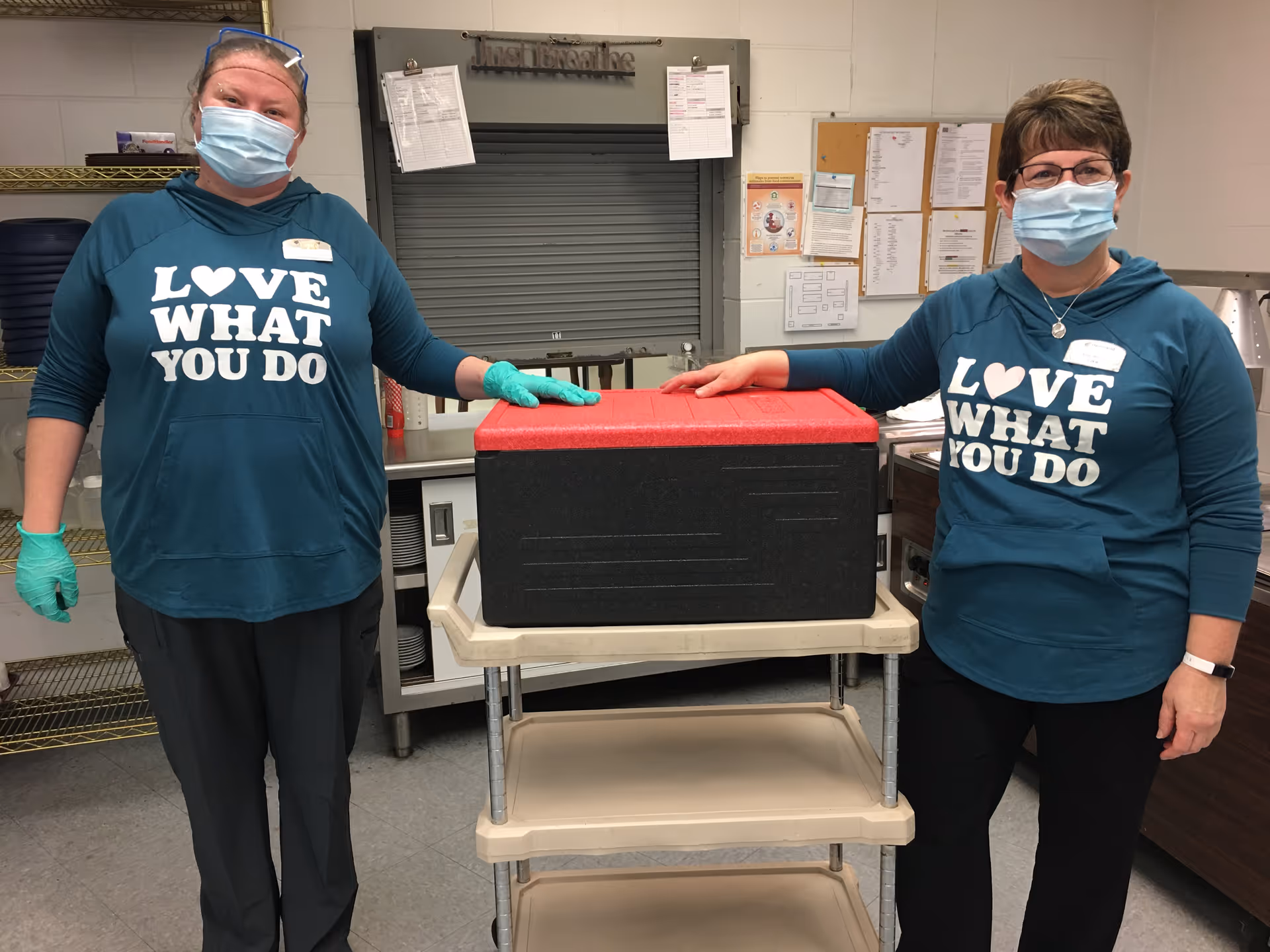 Two women wearing teal hoodies with the text 'LOVE WHAT YOU DO', face masks, and gloves stand on either side of a beige utility cart with a large black and red insulated container on top in a kitchen or food service area.