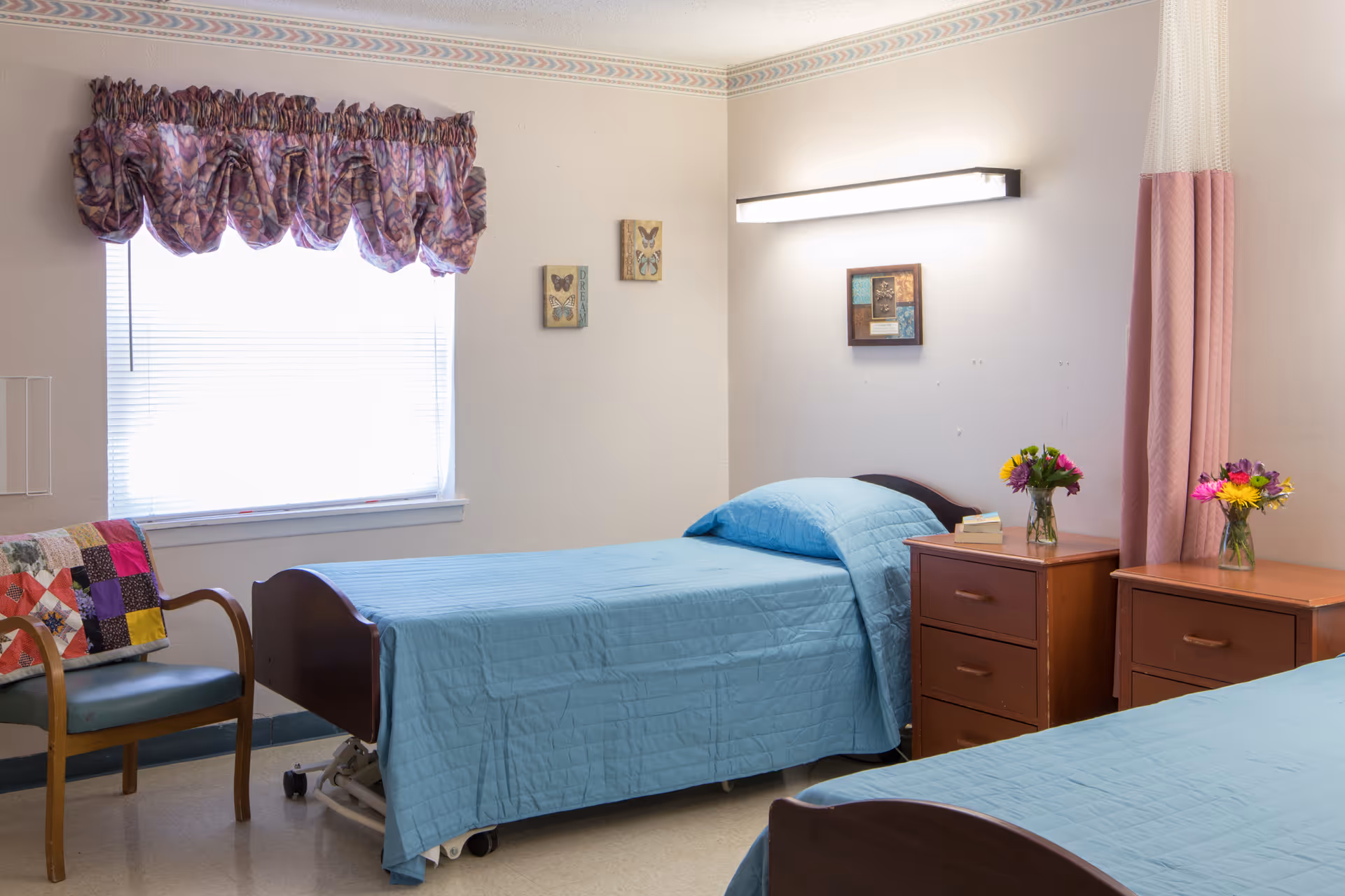A nursing facility room with two single beds covered in light blue bedspreads. Each bed has a wooden nightstand next to it, with a vase of colorful flowers on top. There is a window with a patterned valance and a chair with a colorful quilt draped over it. The walls are light-colored with small framed butterfly artwork and a wall-mounted light above one bed.